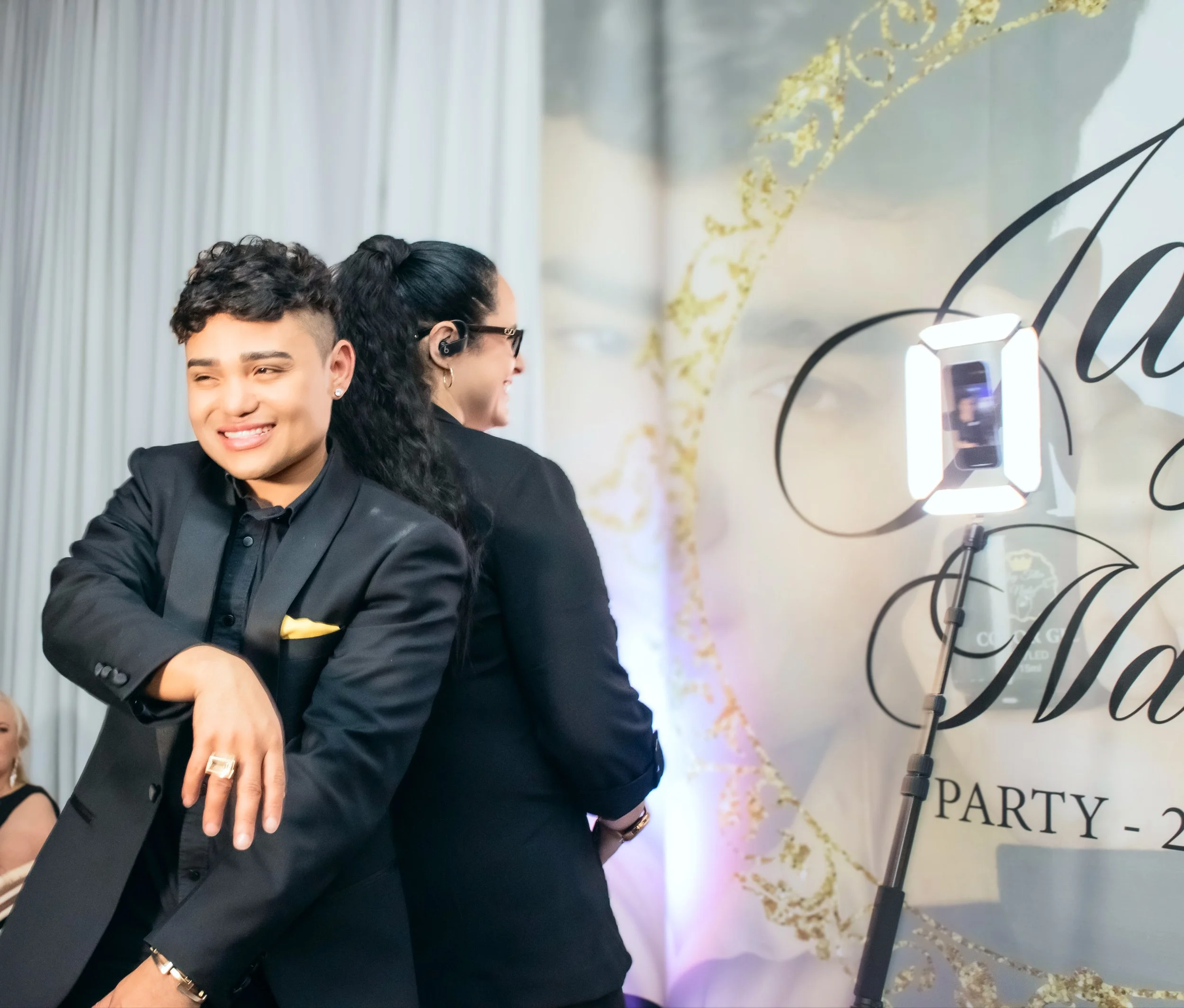 Two people standing at a party, one smiling with arms crossed and the other facing the side near a photo booth.