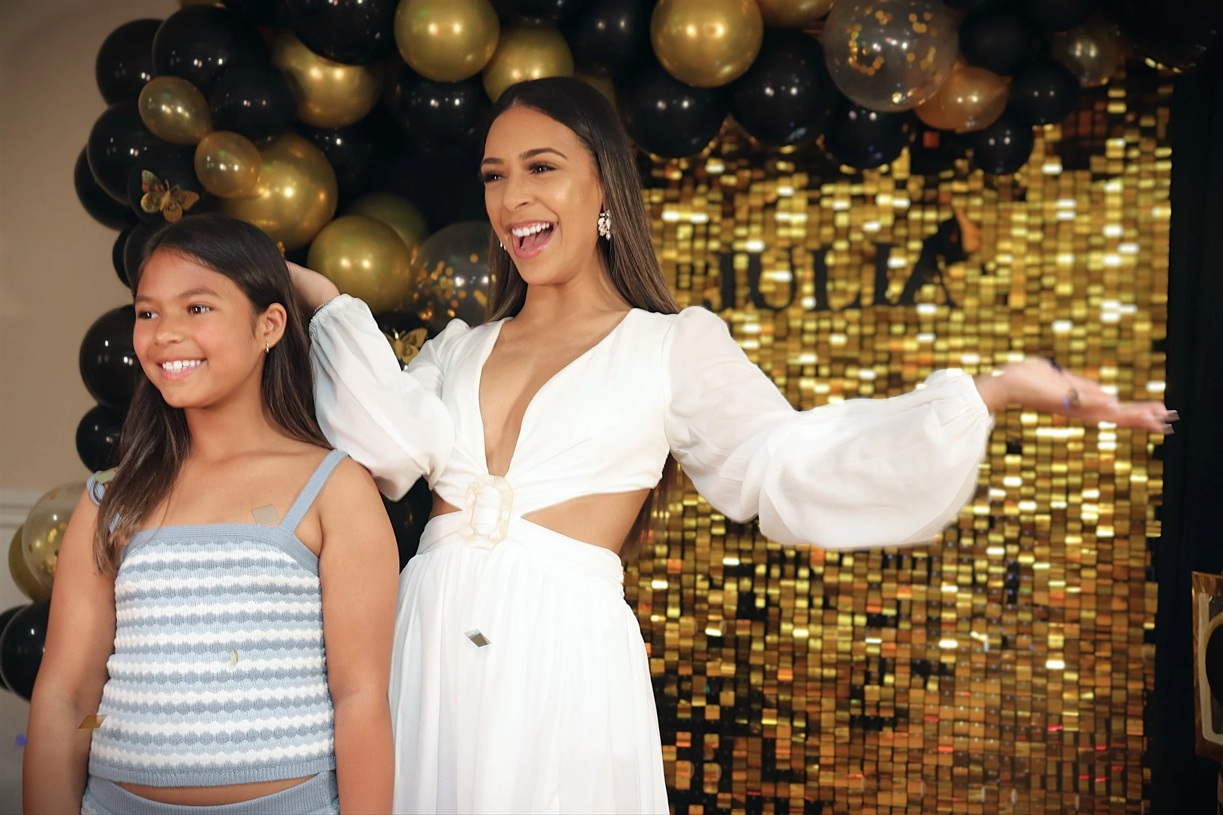 A woman and a girl standing together at a celebration with black and gold balloons and a gold sequin backdrop. They are smiling and enjoying the moment.