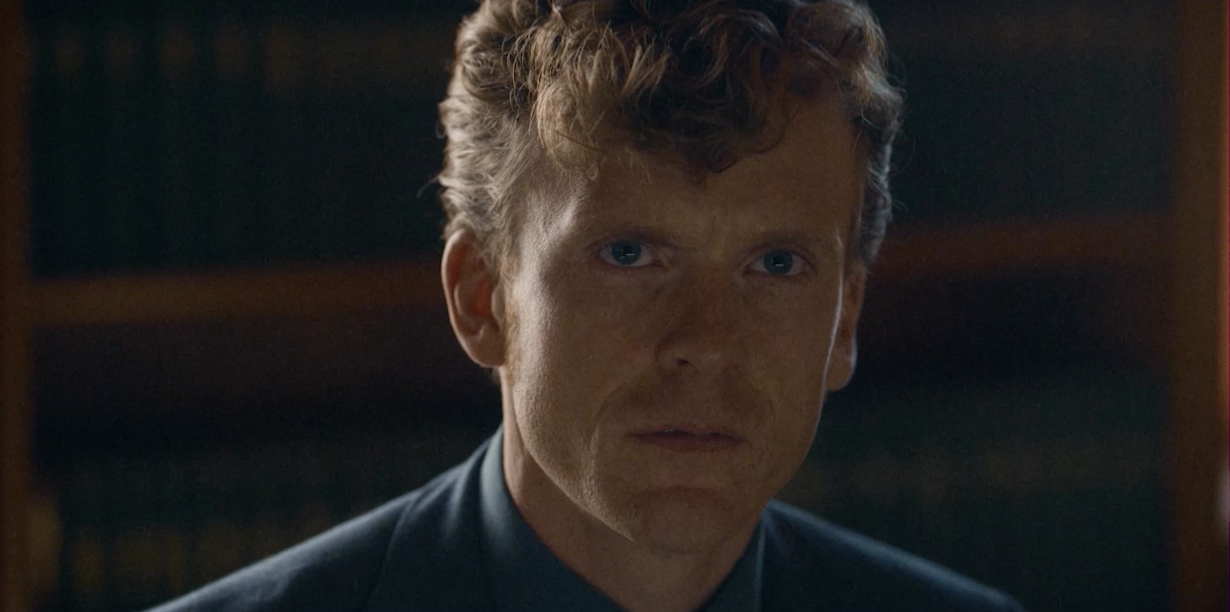 Close-up of a young man with curly brown hair and blue eyes, wearing a dark suit, in a dimly lit room.