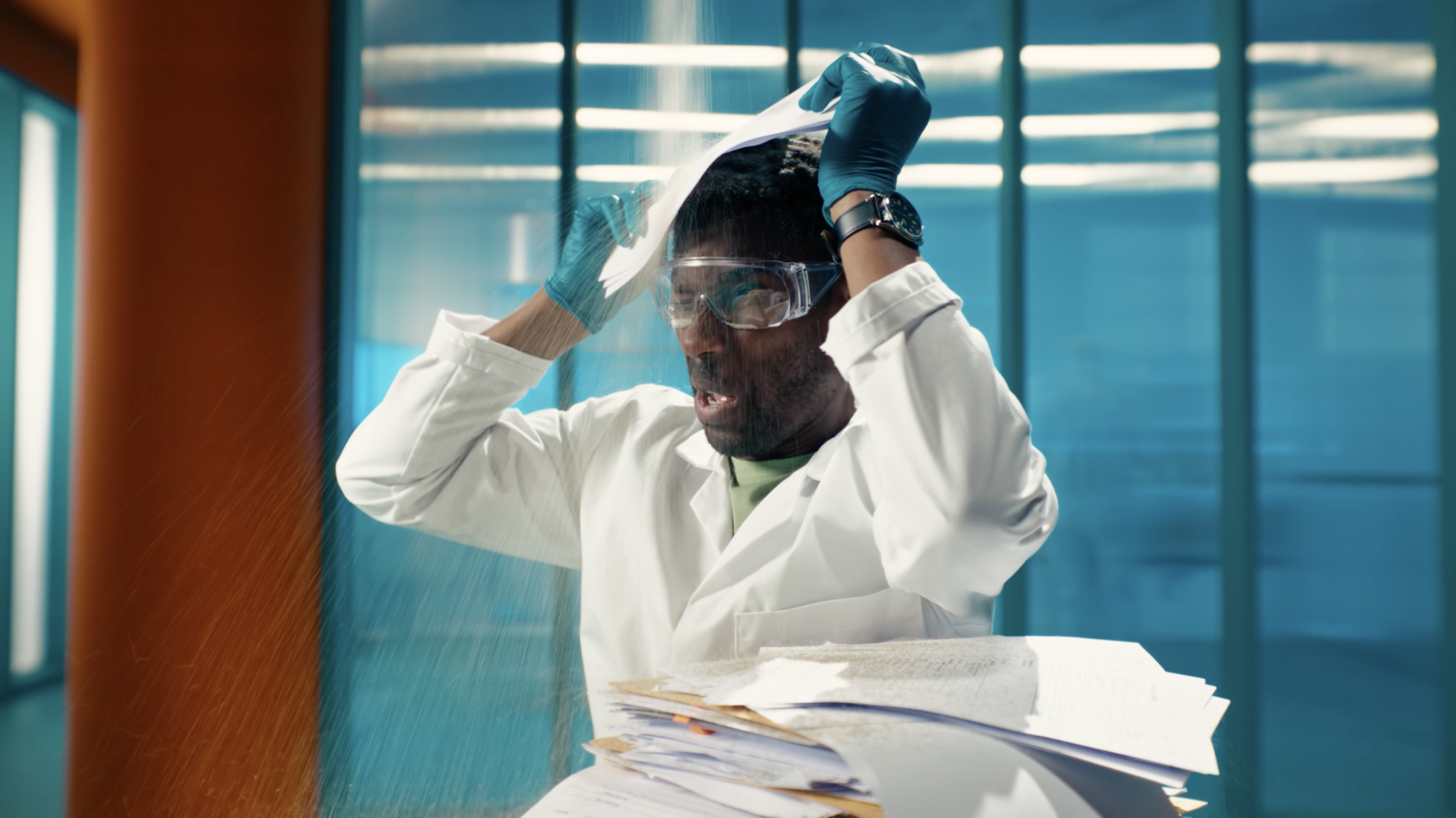 A scientist wearing safety goggles, gloves, and a white lab coat adjusts a face shield in a laboratory with blue and orange panels and stacks of papers on the table.