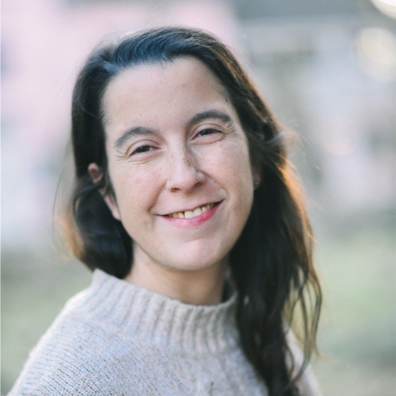 A woman with straight, shoulder-length dark hair, fair skin, and blue eyes, smiling for a portrait with a blurred warm-toned background, wearing a white top.
