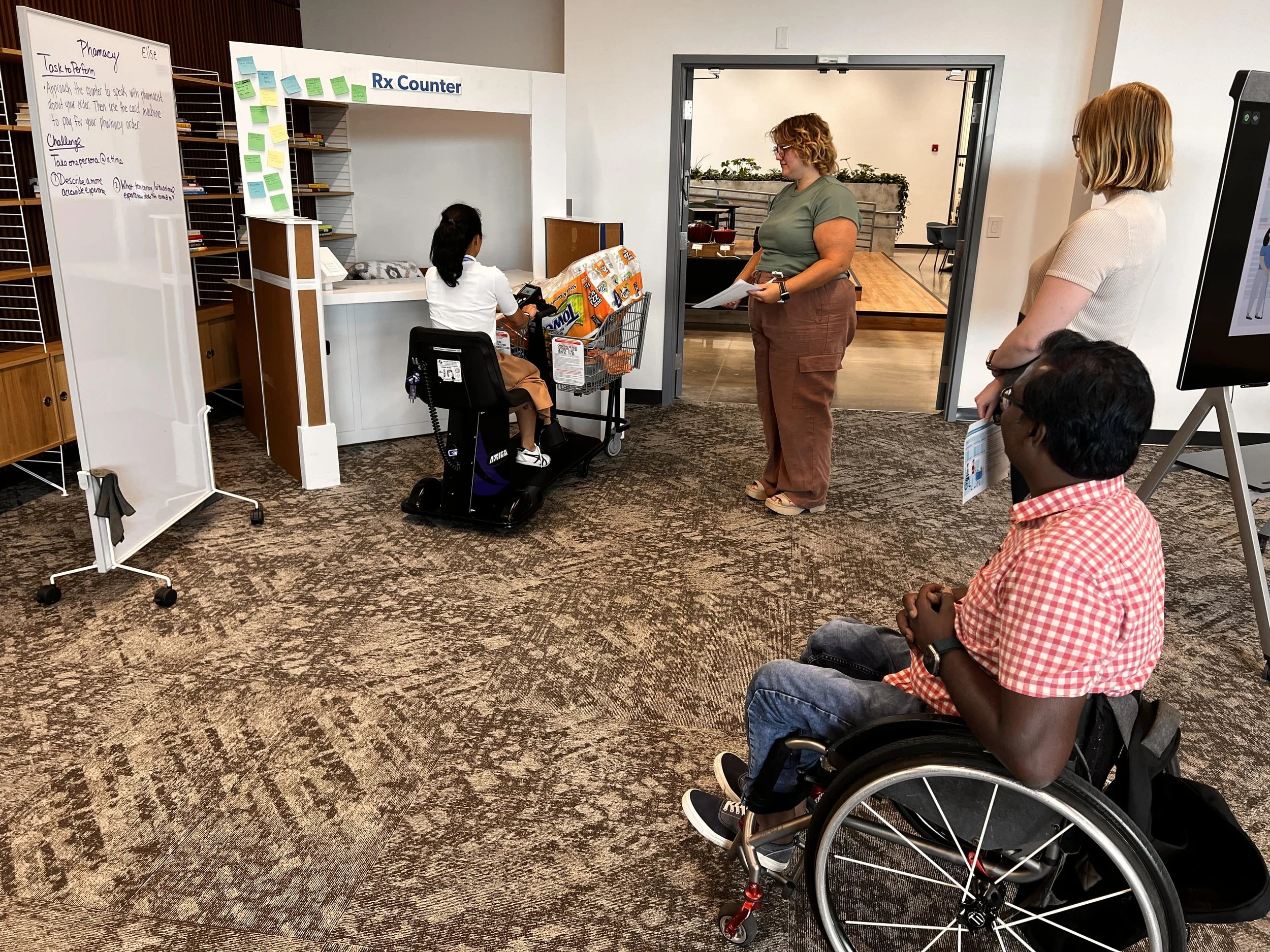 Workshop participants using a low-fidelity foam-core prototype of an Rx counter to experience accessing the counter from a wheelchair, with others observing and discussing the interaction.