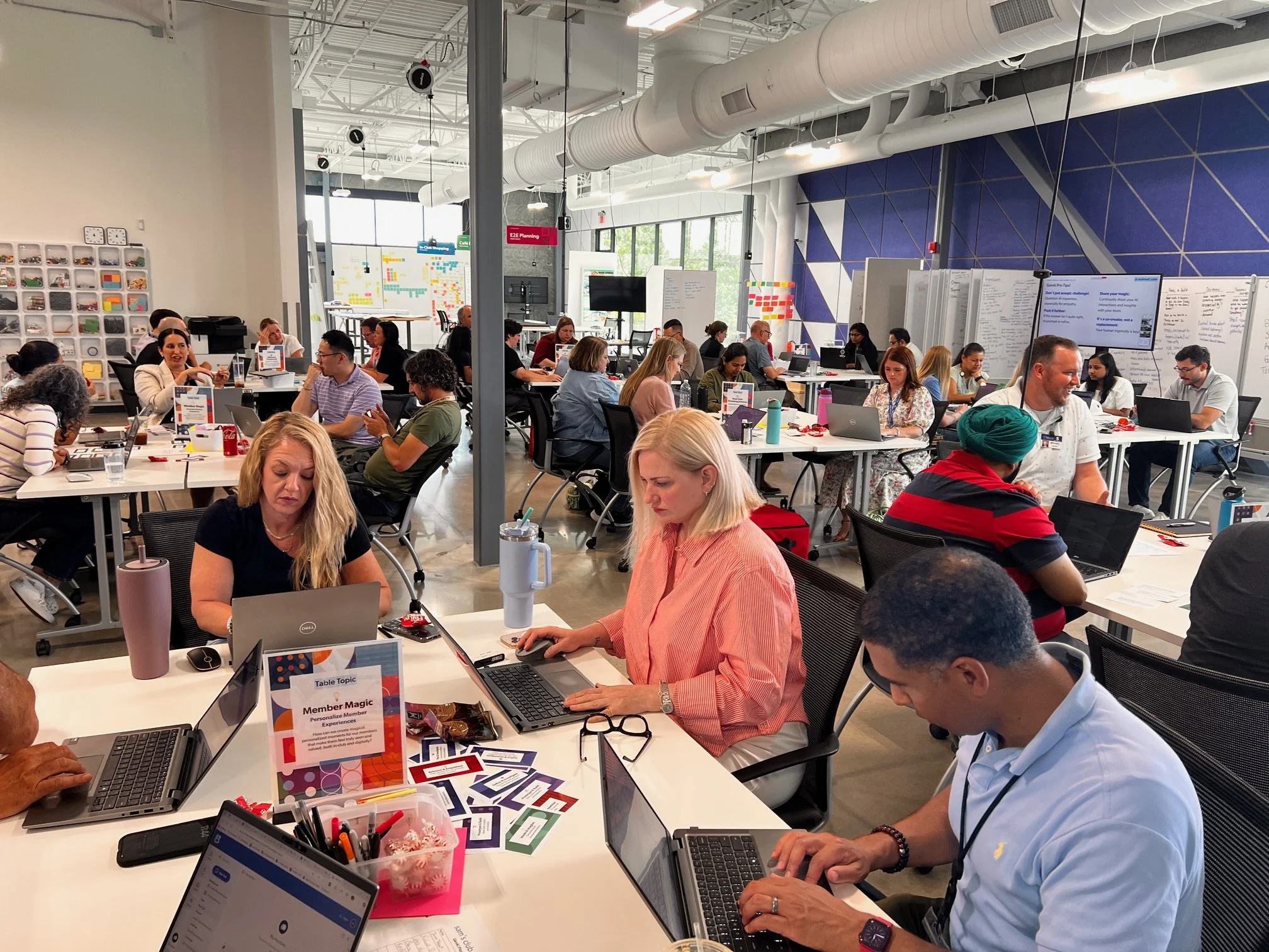 Groups of people collaborating at tables in a large workshop space, working together on laptops and materials.