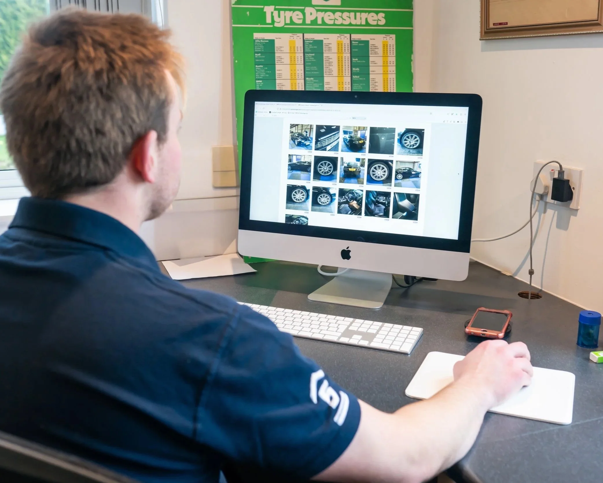 A person sitting at a desk looking at photos of a car storage check-in report on an iMac computer screen.