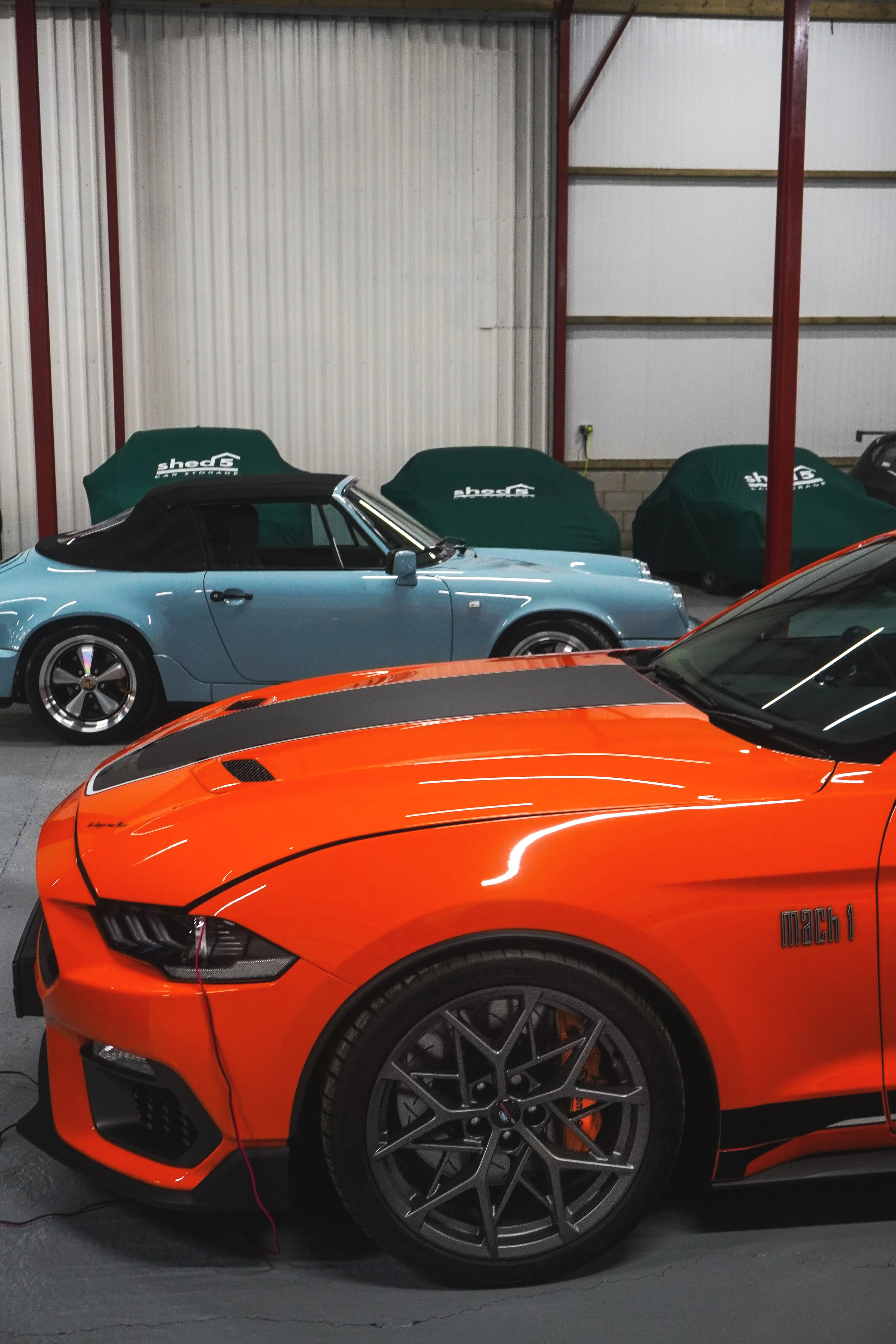 inside a white building ,an orange ford mustang is in the foreground with a blue vintage Porsche parked behind it. at the back is a row of cars covered in matching green covers  