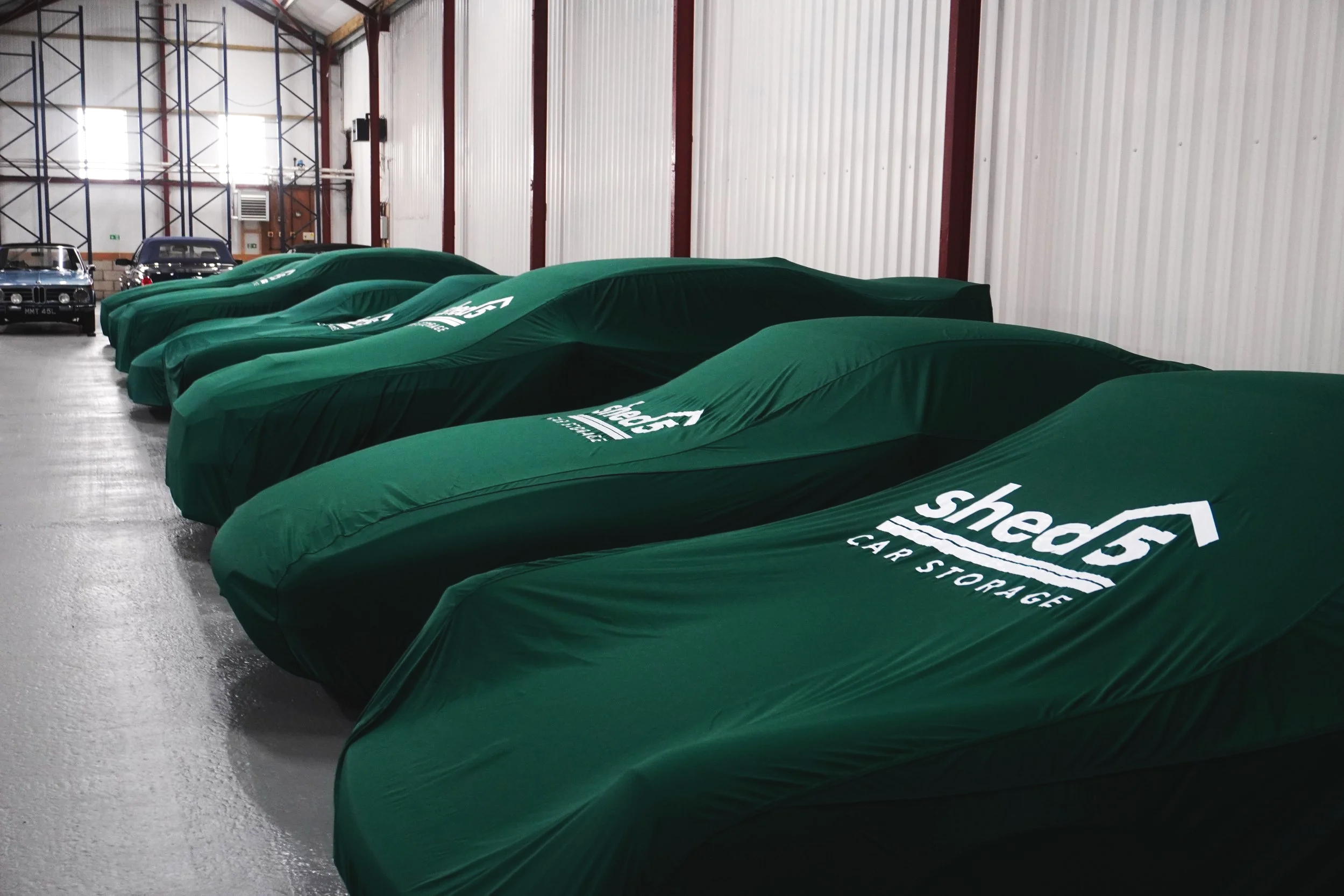 7 cars in a row inside a building with GREEN CAR COVERS WITH THE LOGO 'SHED 5 CAR STORAGE'