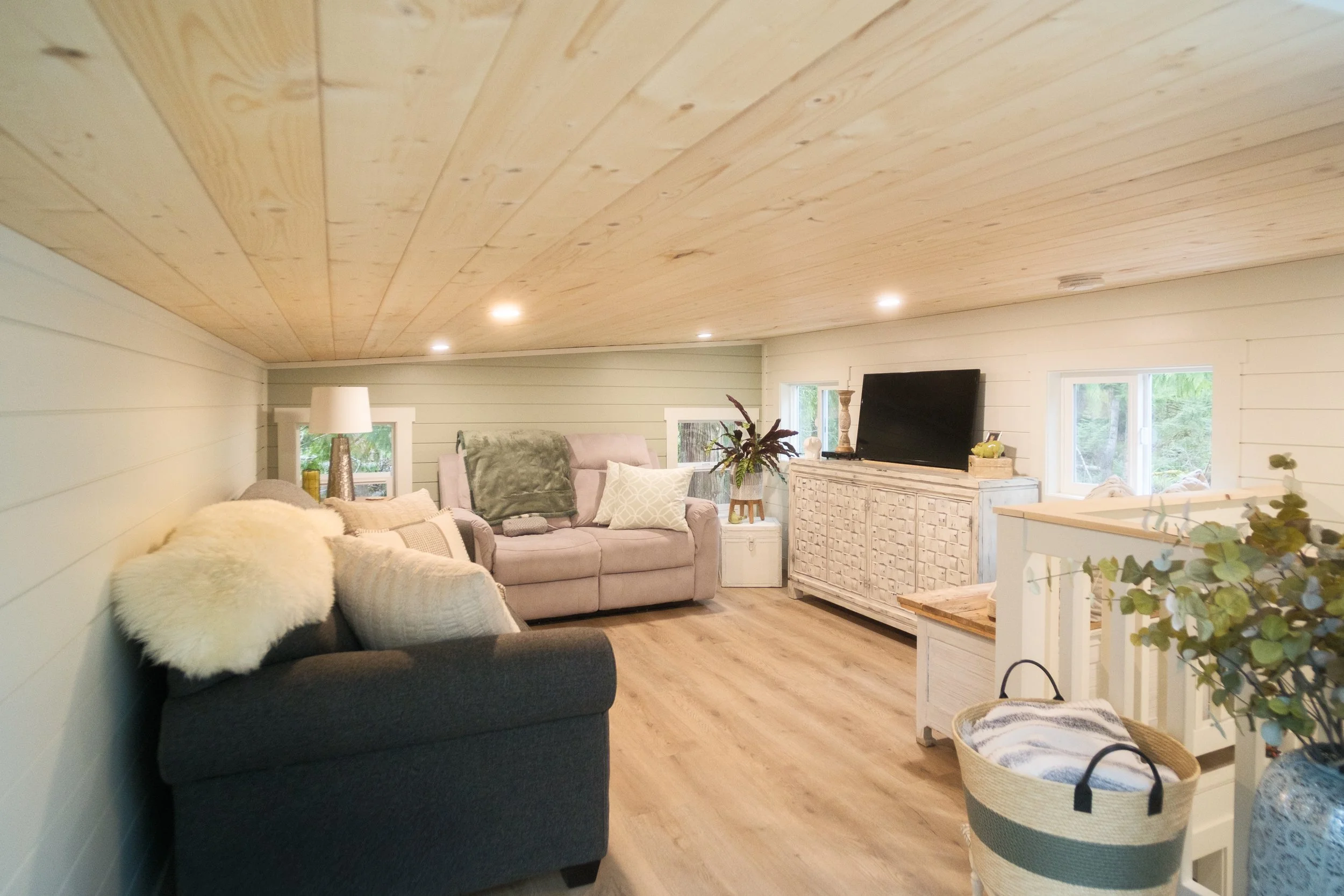 Cozy loft with a sloped wooden ceiling, light-colored walls, hardwood flooring, a black couch with a white furry throw, a pink armchair, a TV on a white textured stand, and various decorative items including plants, lamps, and small furniture. Dwell 