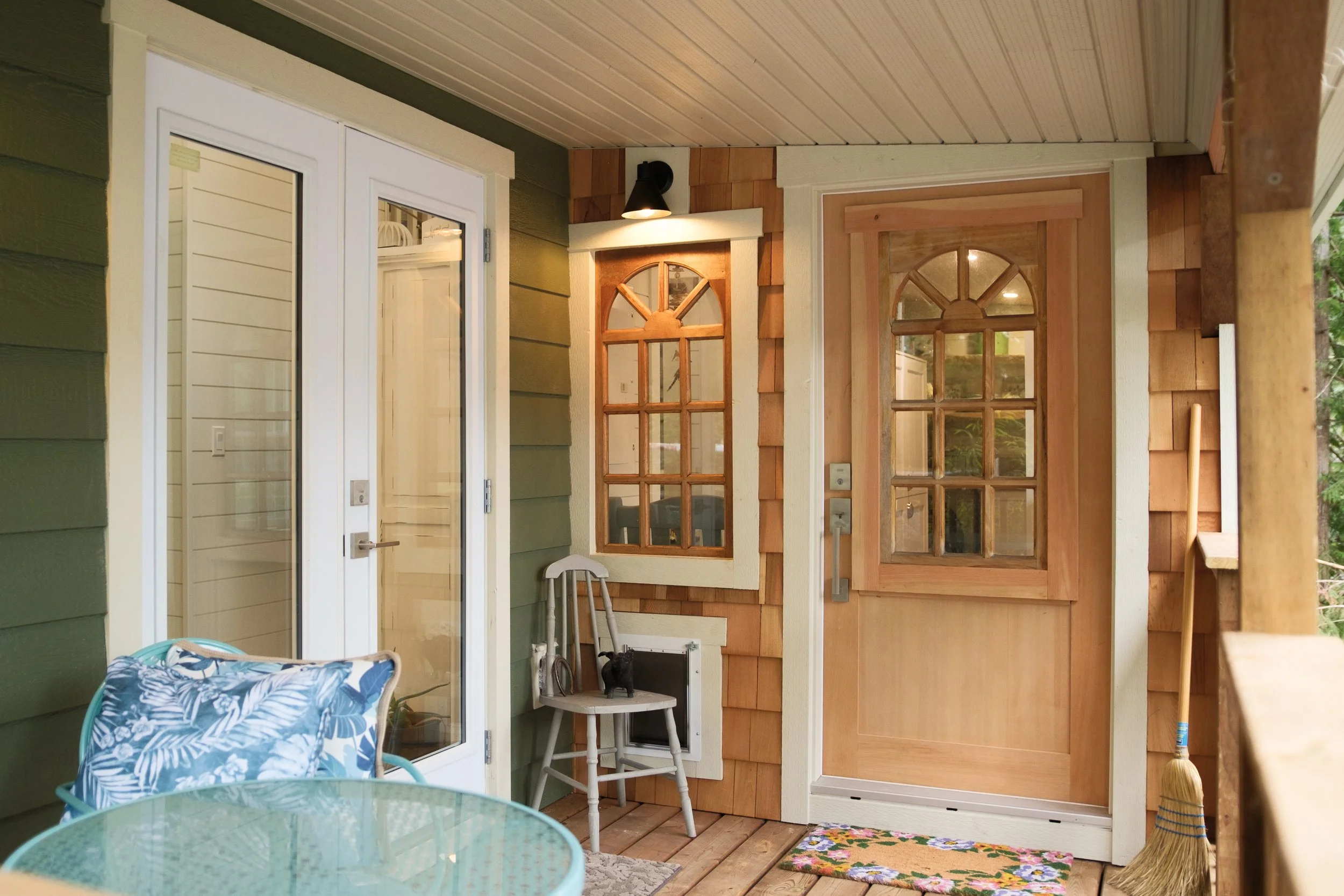 Dwell Tiny Homes 400sq. ft. model. A front porch of the Dwell Tiny Homes 400 sq. ft.  model with a wood door, a glass door, a chair with a black cat, and a broom leaning against the wall.