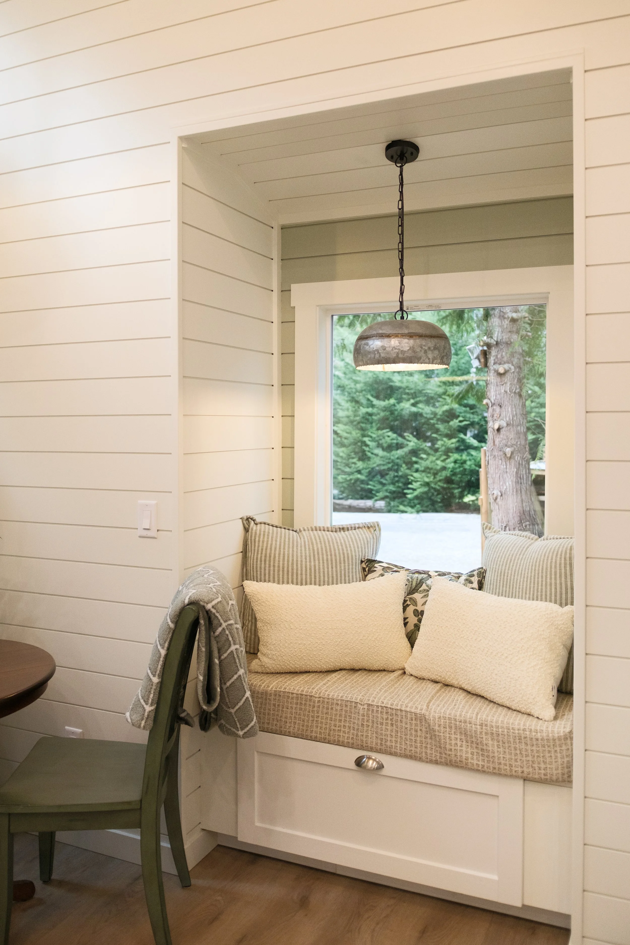 A cozy window nook with a cushioned seat, four decorative pillows, and a view of trees outside, illuminated by a hanging pendant light. Dwell Tiny Homes 400sq. ft. model. Parksville, B.C. Canada