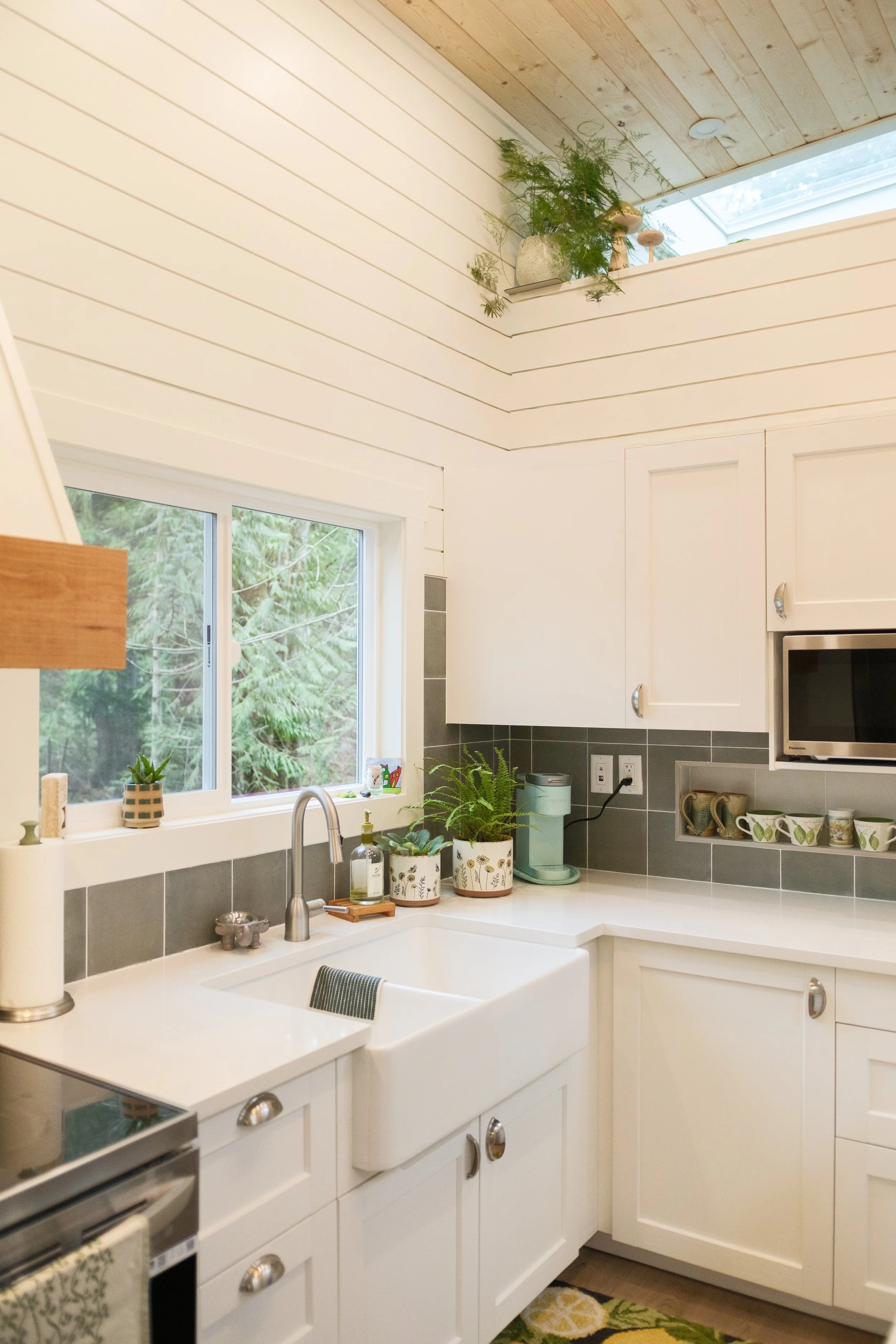 Cozy kitchen with white cabinets, a farmhouse sink, a window overlooking trees, and decorative plants on the countertop and high shelf. Dwell Tiny Homes 400sq. ft. model. Parksville, B.C. Canada