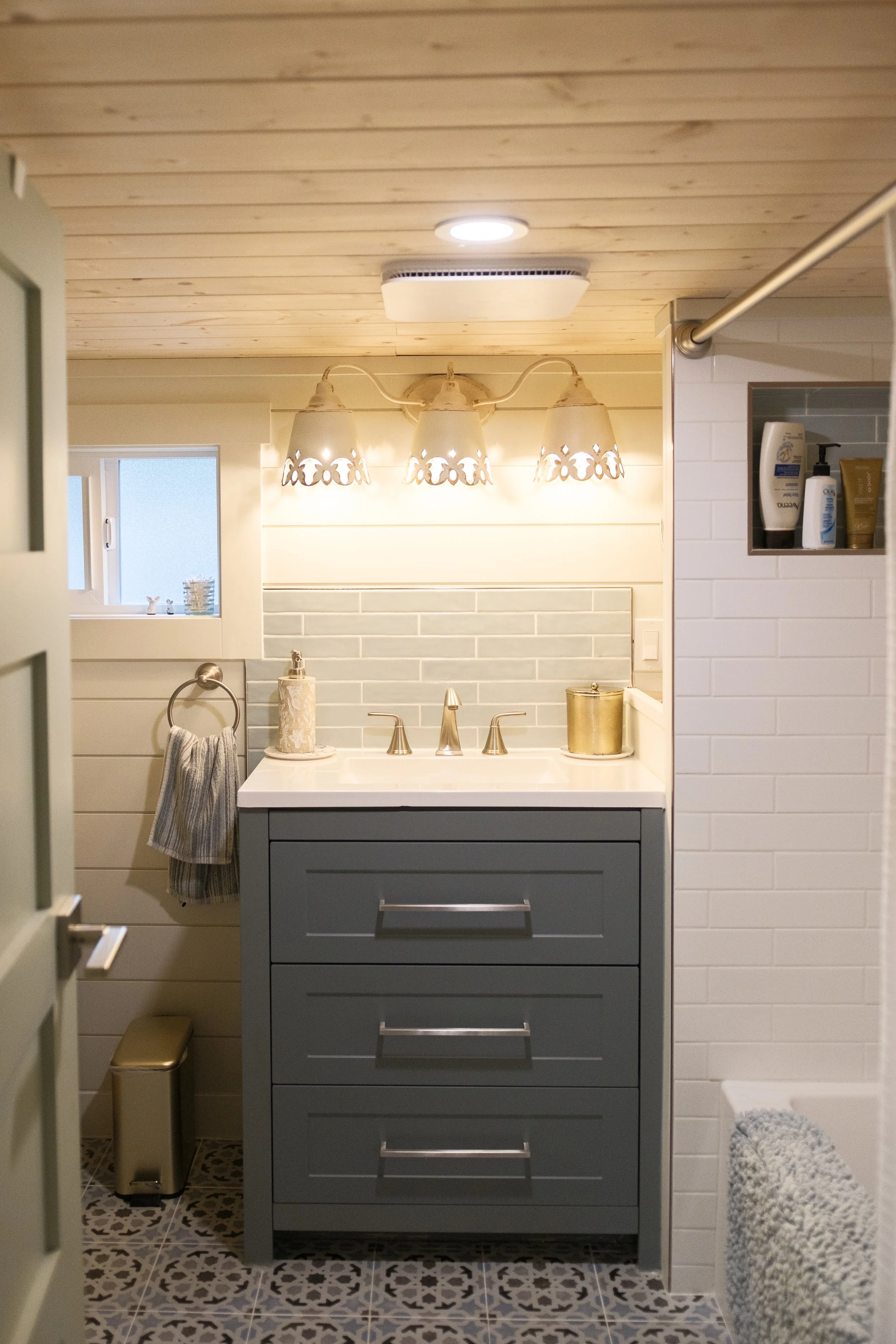 A bathroom vanity with a gray cabinet, a white sink, and brass fixtures, illuminated by a four-lamp chandelier, with a small window and tiled wall in the background. Dwell Tiny Homes 400sq. ft. model. Parksville, B.C. Canada