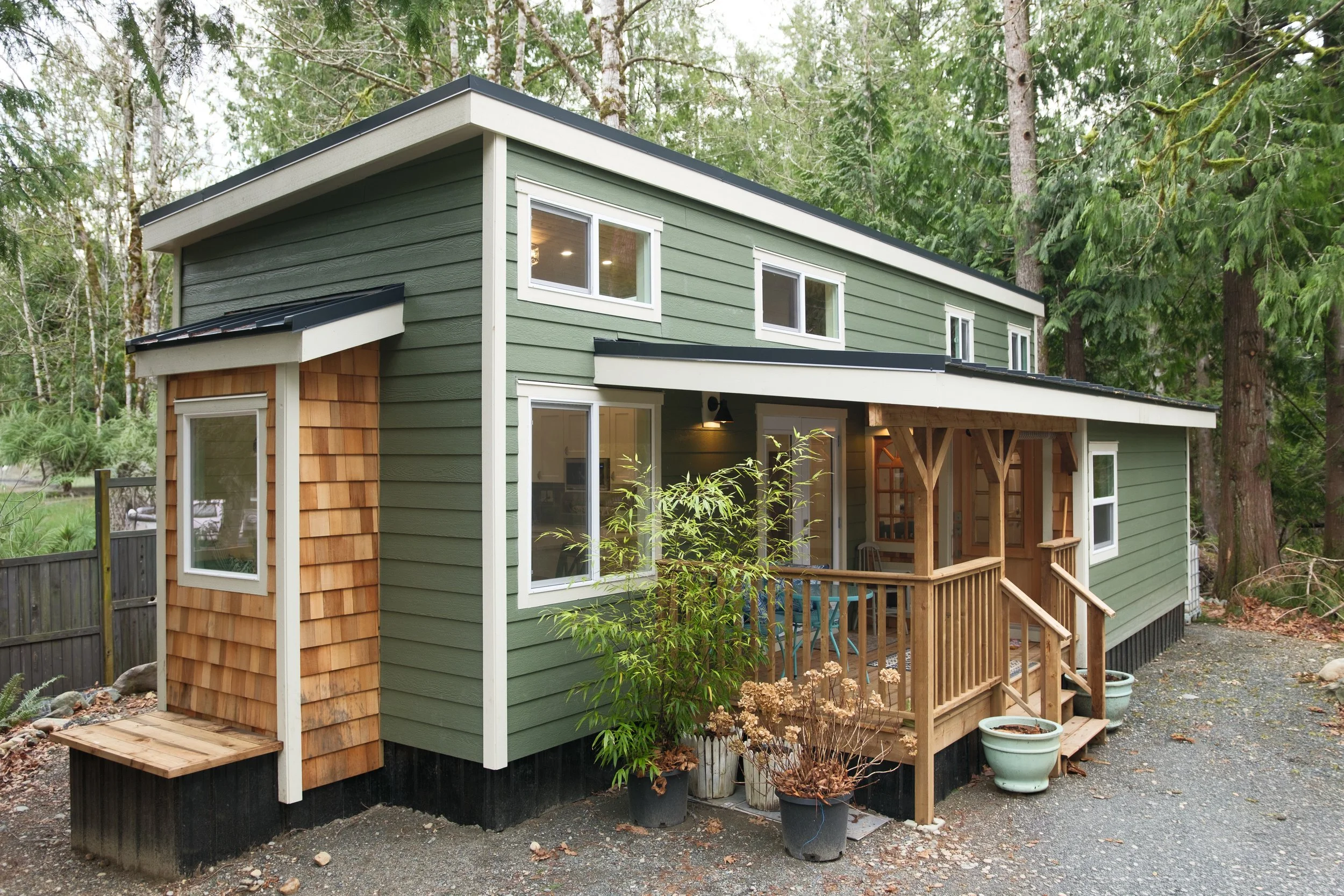 Dwell Tiny Homes 400sq. ft. model. Green tiny home with white trim, small porch with wooden railing, surrounded by trees and potted plants. Delivered to Qualicum Beach, Vancouver Island. 