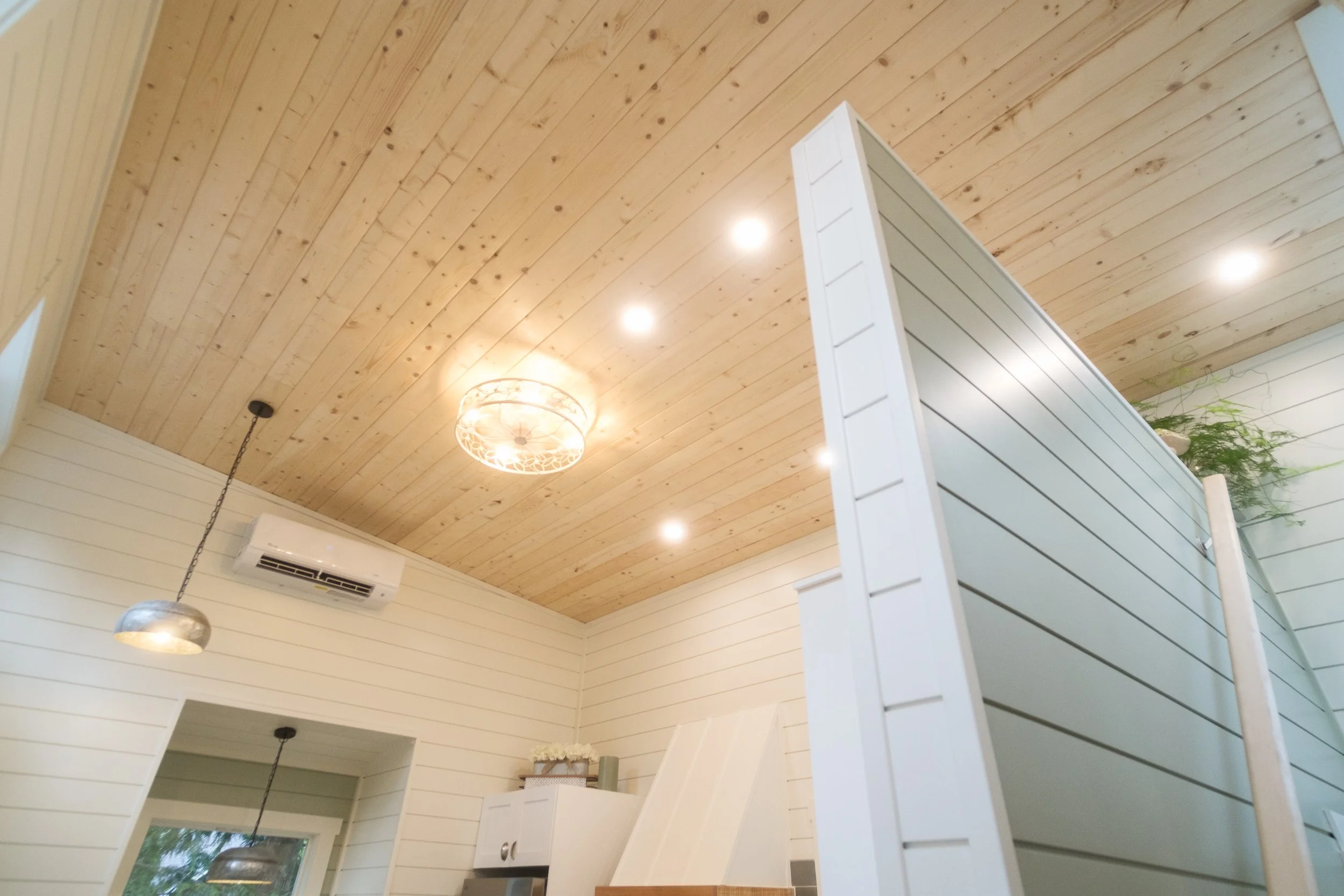 Interior of a room with light wood ceiling, white walls with horizontal paneling, and a white partition wall. There are ceiling lights, a hanging pendant light, an air conditioning unit, and some decorative items on a cabinet, with a window showing g