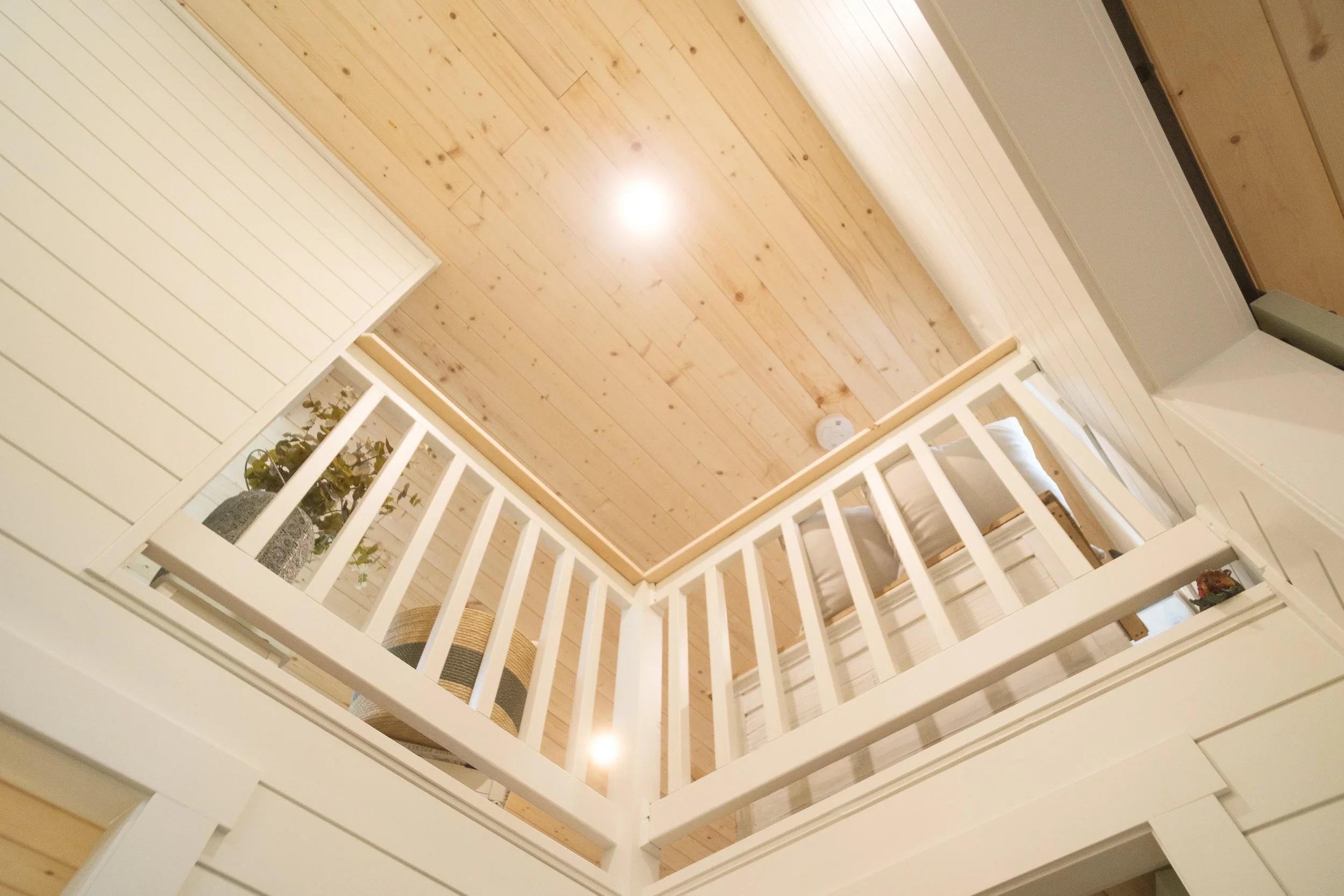 Interior view looking up at a wooden ceiling and a white railing surrounding an open space with a bed and decorative plant. Dwell Tiny Homes 400sq. ft. model. Parksville, B.C. Canada