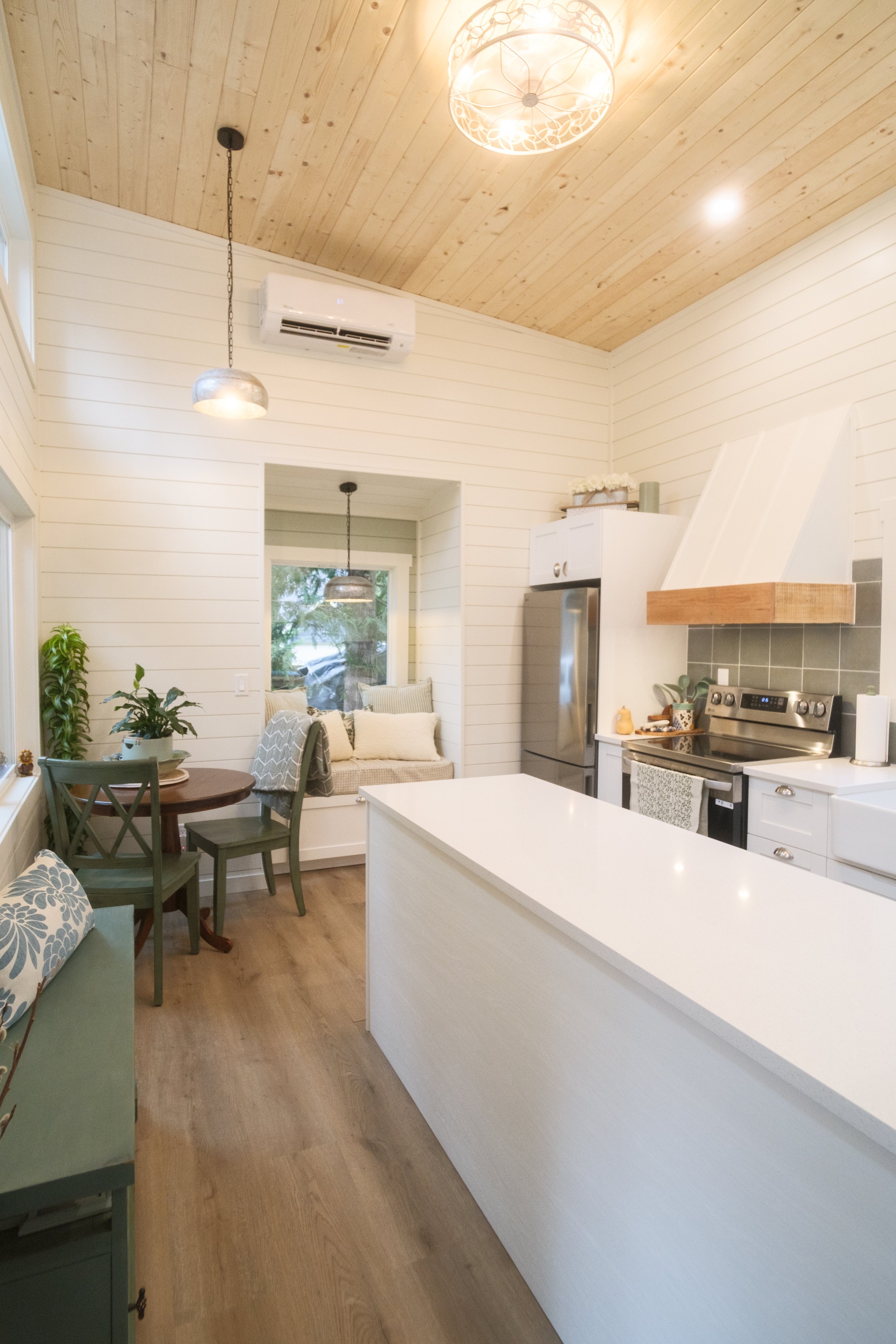 Interior view of a bright, cozy kitchen with white shiplap walls, a wooden ceiling, a kitchen island with a white countertop, stainless steel appliances, a small nook with a window seat and cushions, a dining table with chairs, and pendant lights.