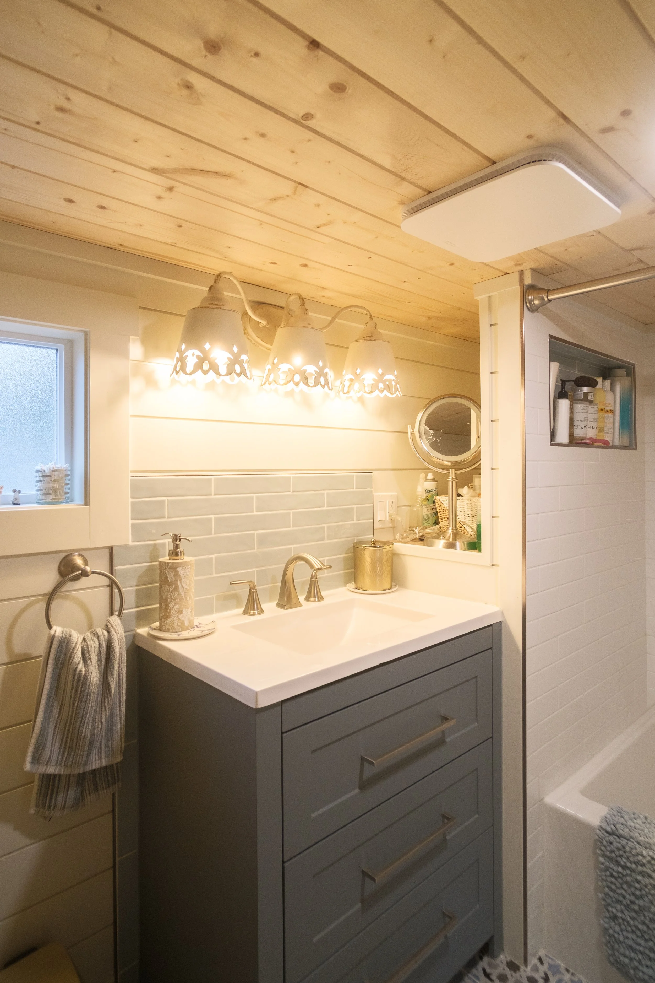 A cozy bathroom with a wooden ceiling, a small window, a vanity with a gray cabinet and white countertop, a round mirror, a soap dispenser, and a gold container. There is a three-light vanity fixture with decorative shades and a white vent on the cei