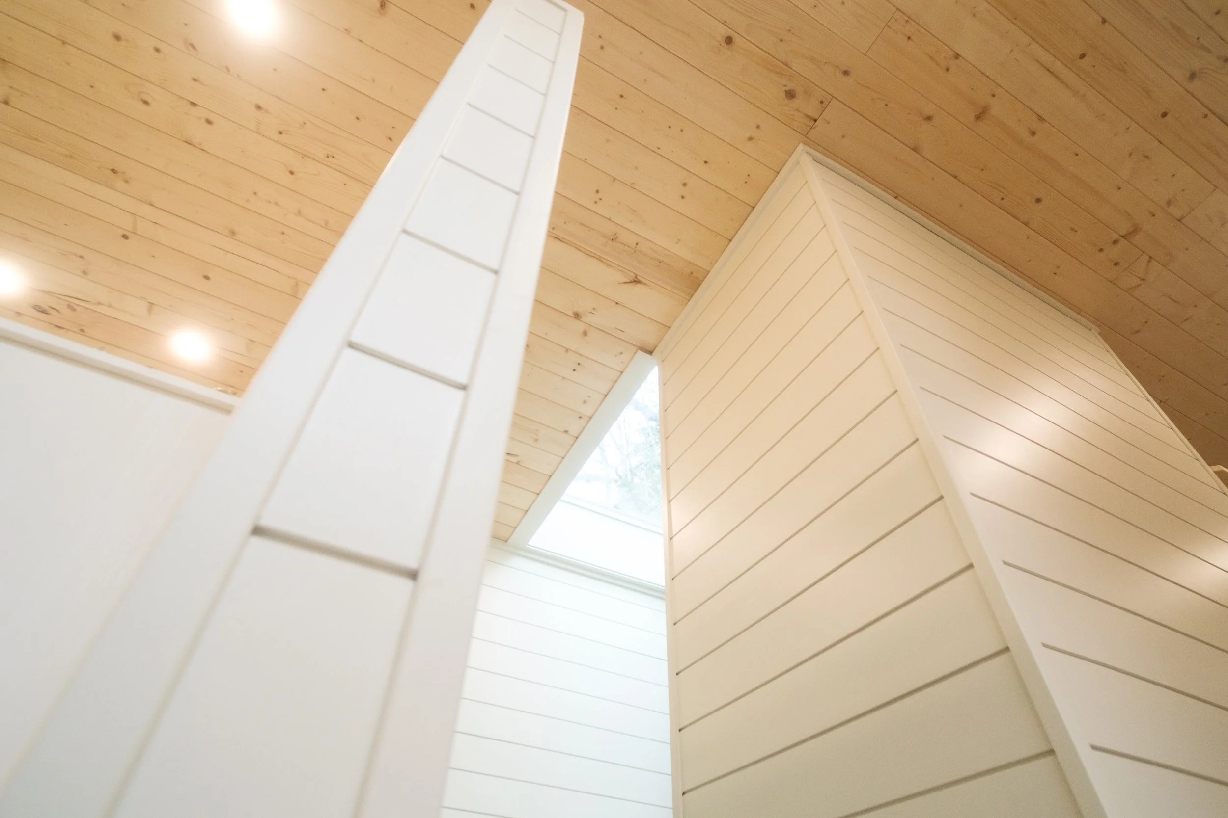 Interior view looking up at a wooden ceiling with natural light through a skylight, and white wooden partition walls with horizontal paneling. Dwell Tiny Homes 400sq. ft. model. Parksville, B.C. Canada