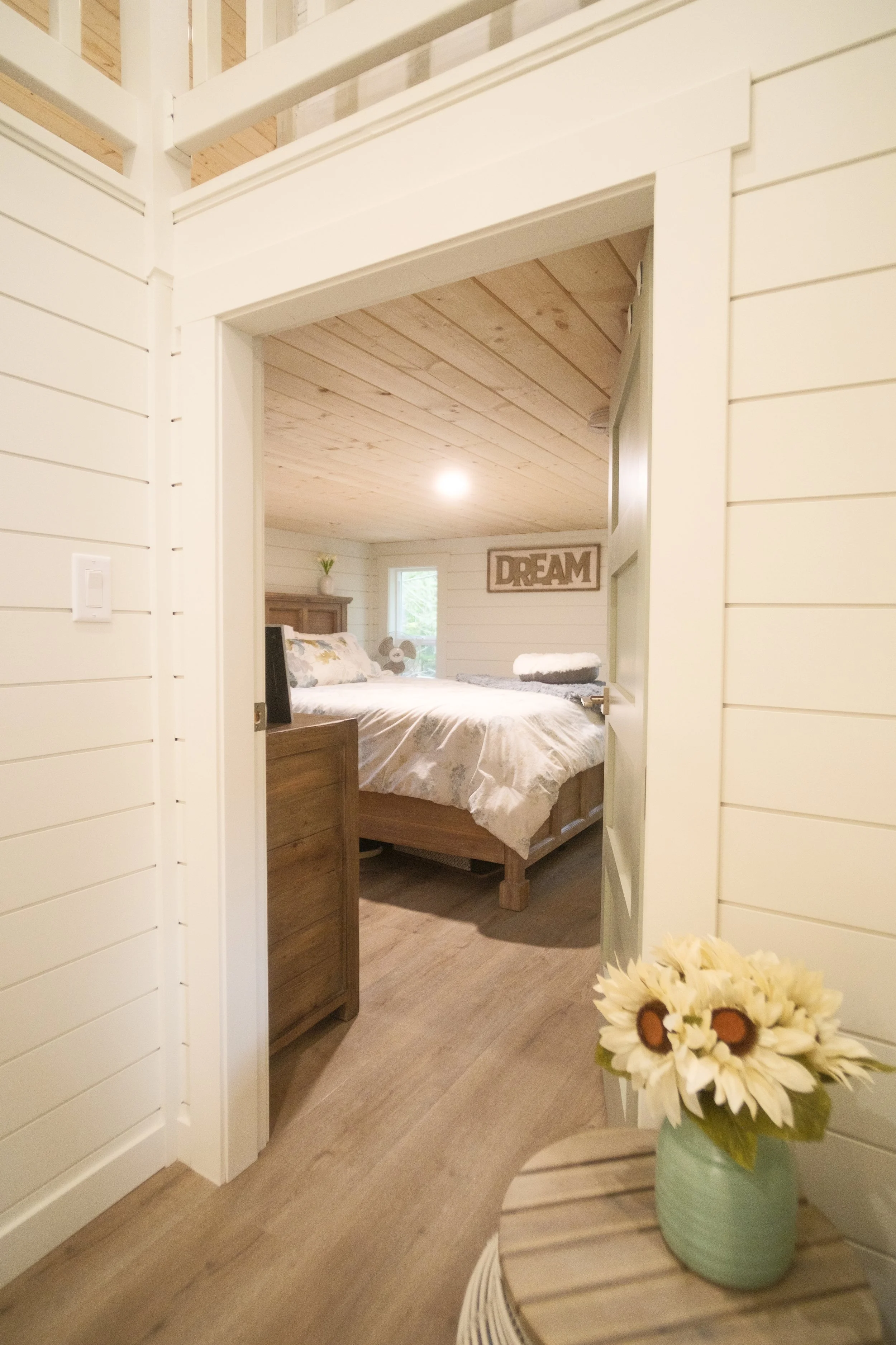 View through an open doorway into a cozy bedroom with a wood-paneled ceiling and a sign that says "DREAM" on the wall. Dwell Tiny Homes 400sq. ft. model. Parksville, B.C. Canada