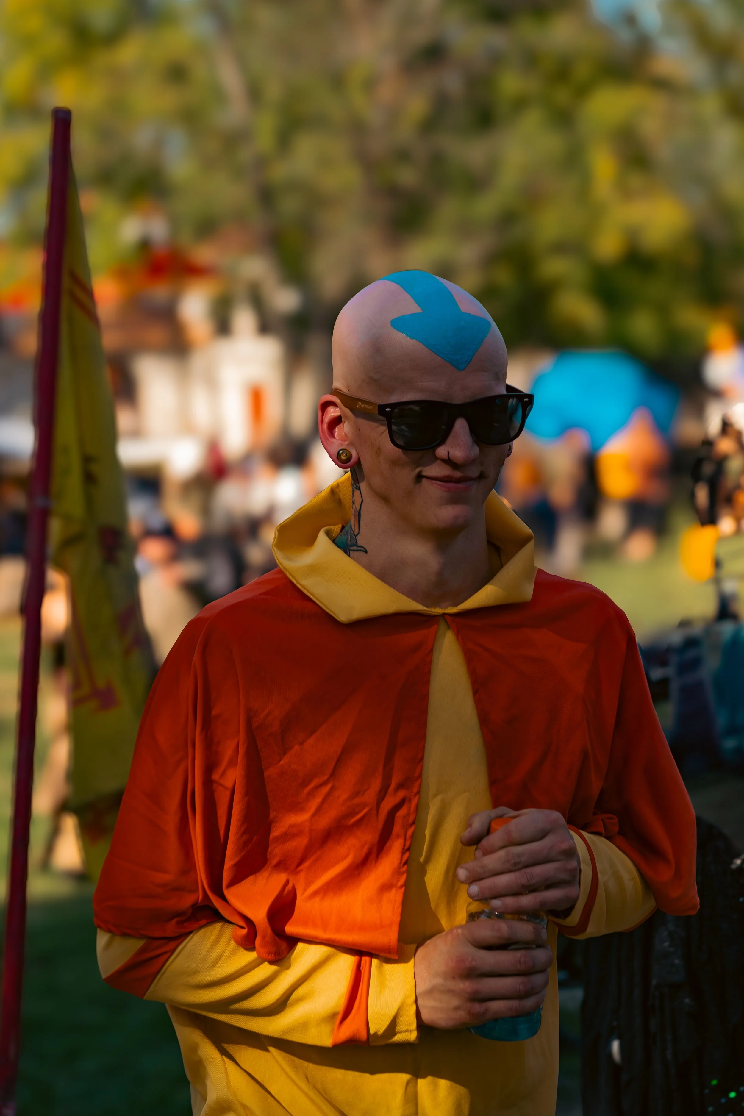 A person with a shaved head, wearing dark sunglasses, yellow and orange jacket, and yellow pants, standing outdoors with a blurred background of trees and people.