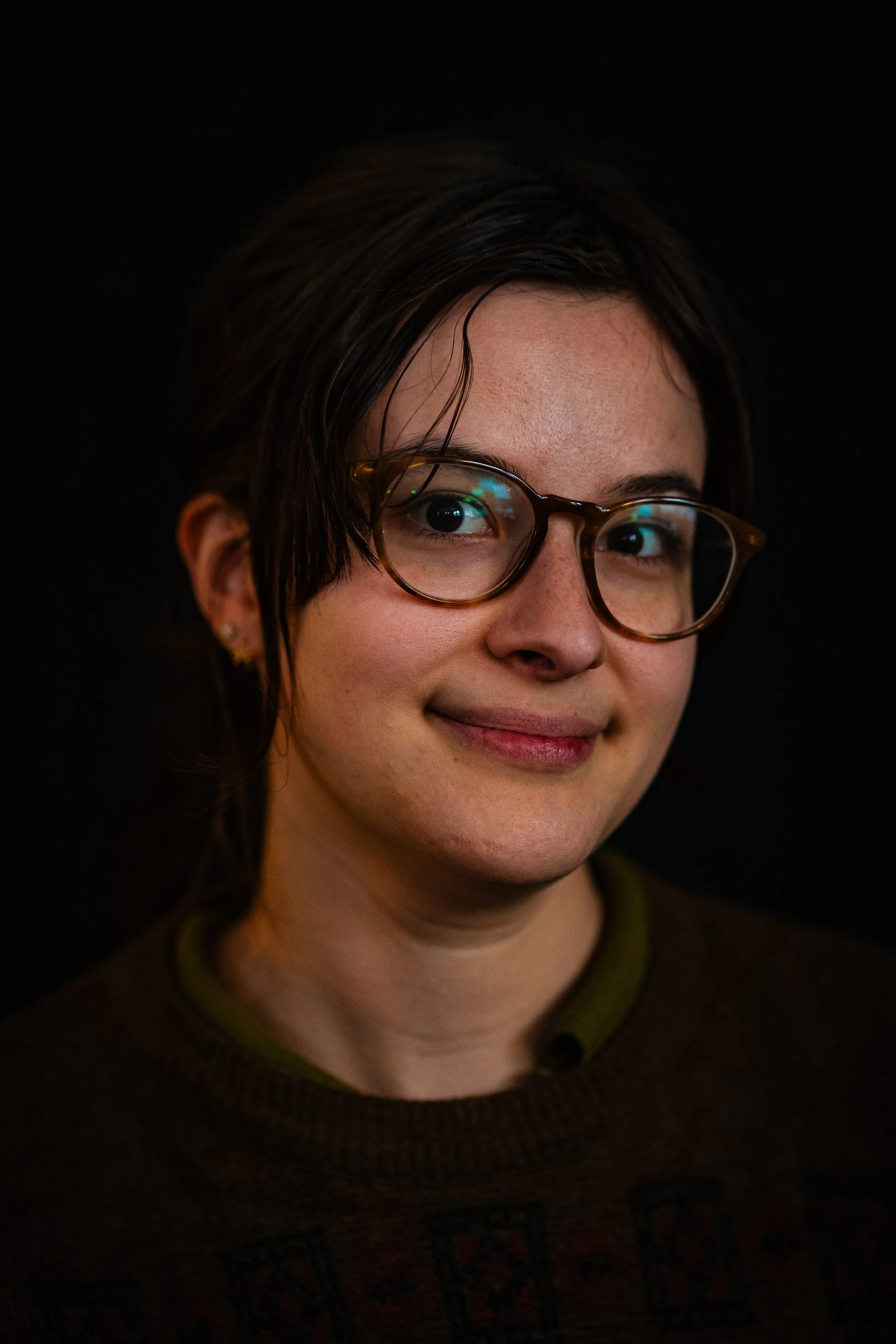 Close-up of a woman with short dark hair, wearing glasses, smiling, against a black background.