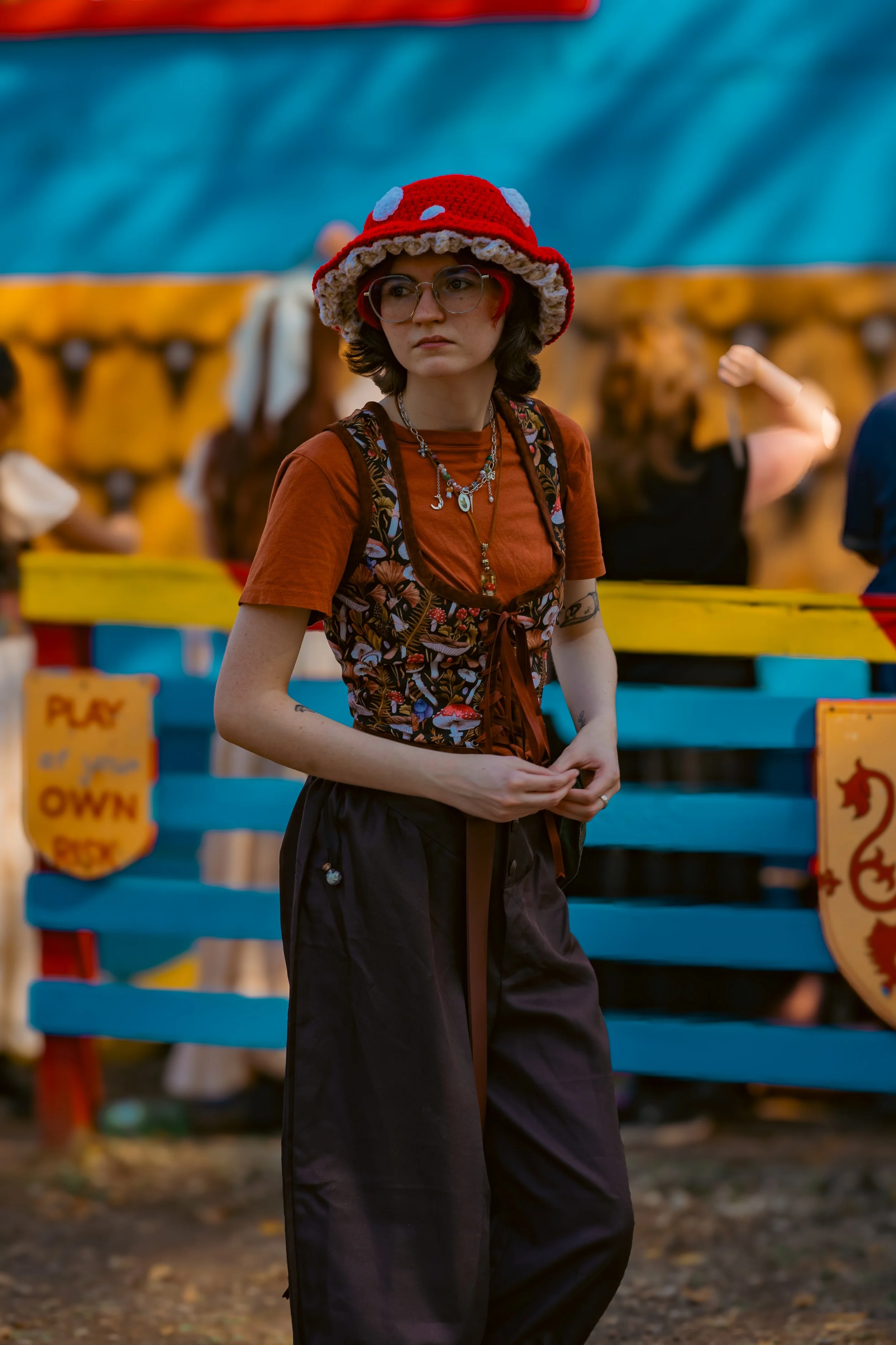 Young woman with glasses, short dark hair, wearing a red crocheted mushroom hat and layered necklaces, standing outdoors in front of colorful carnival booths.