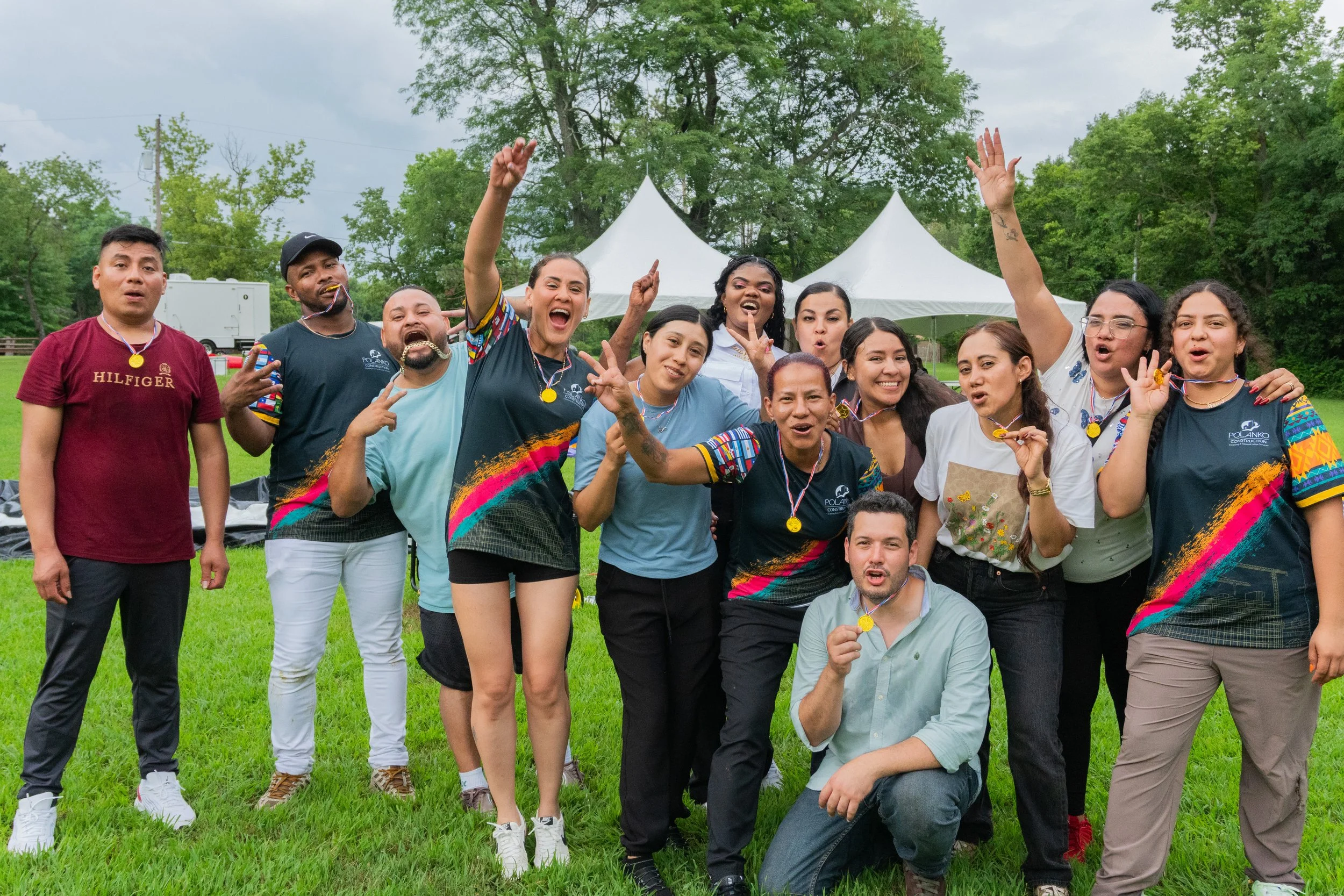Group of diverse people celebrating outdoors with medals, some making peace signs and posing happily on a grassy field with tents and trees in the background.
