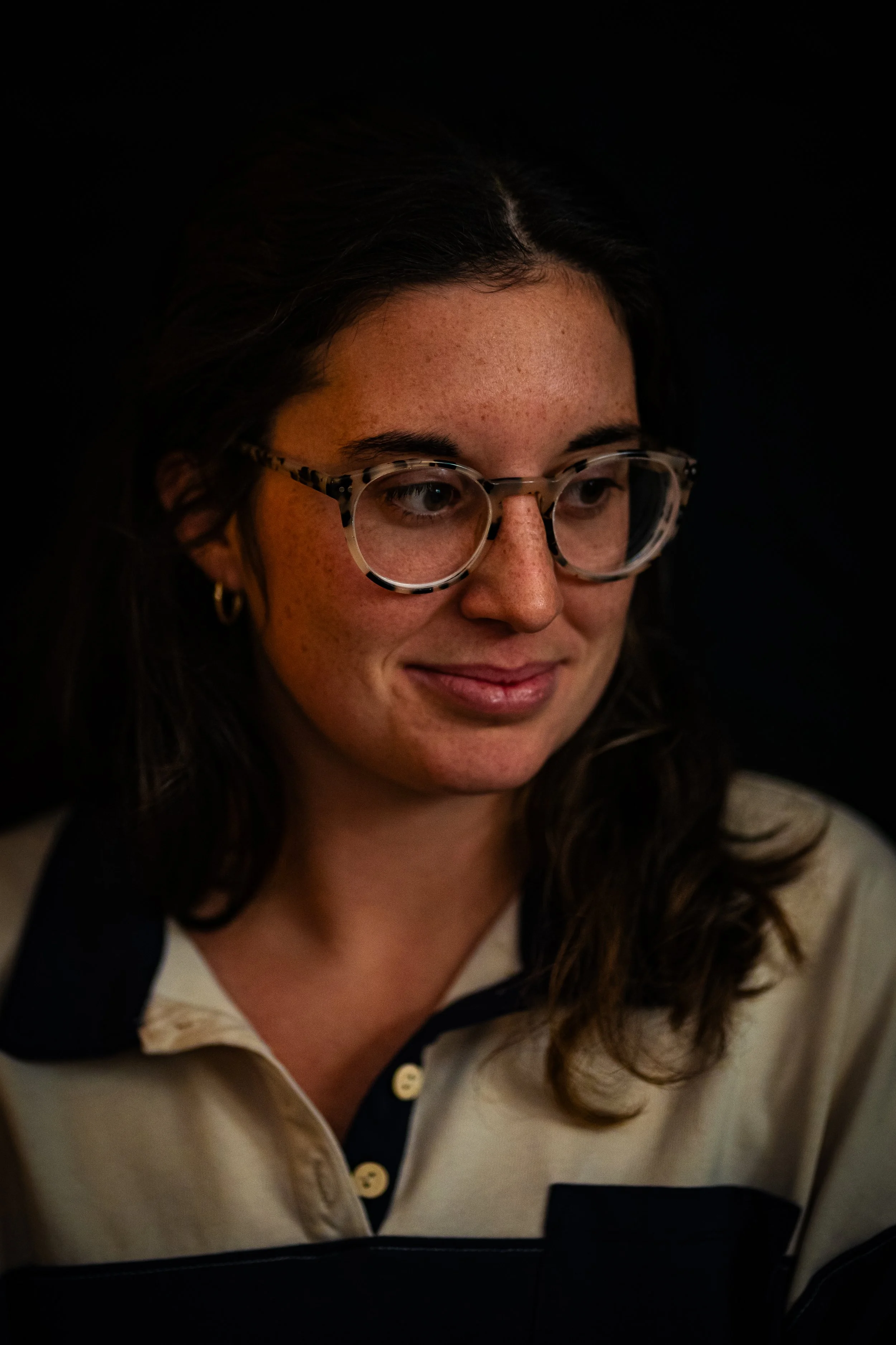 Close-up of a woman with dark hair, wearing large tortoiseshell eyeglasses and a beige and navy shirt, smiling softly with a black background.