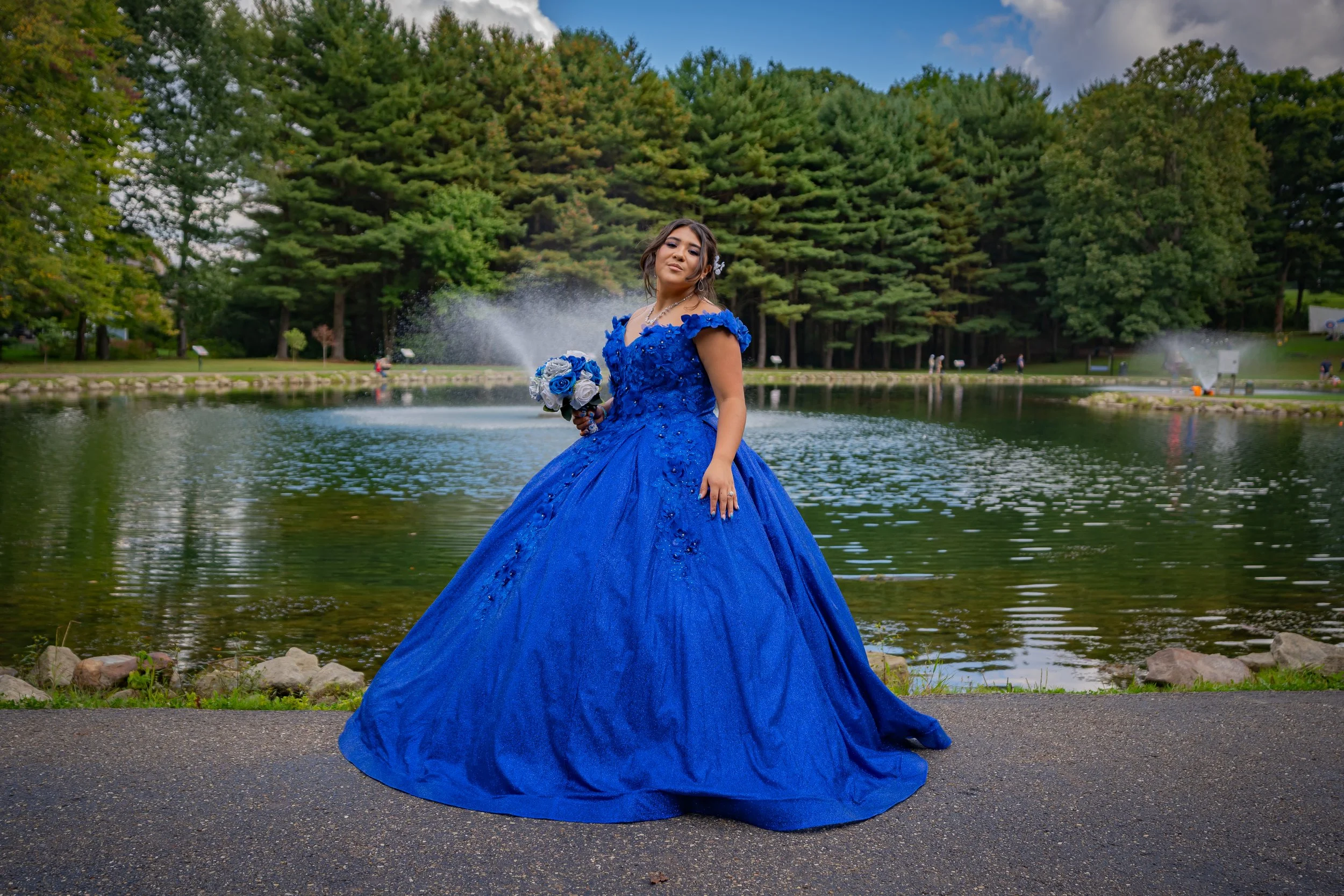 A woman standing near a pond wearing a vibrant blue gown with intricate details, holding a matching bouquet, with water fountains and green trees in the background.
