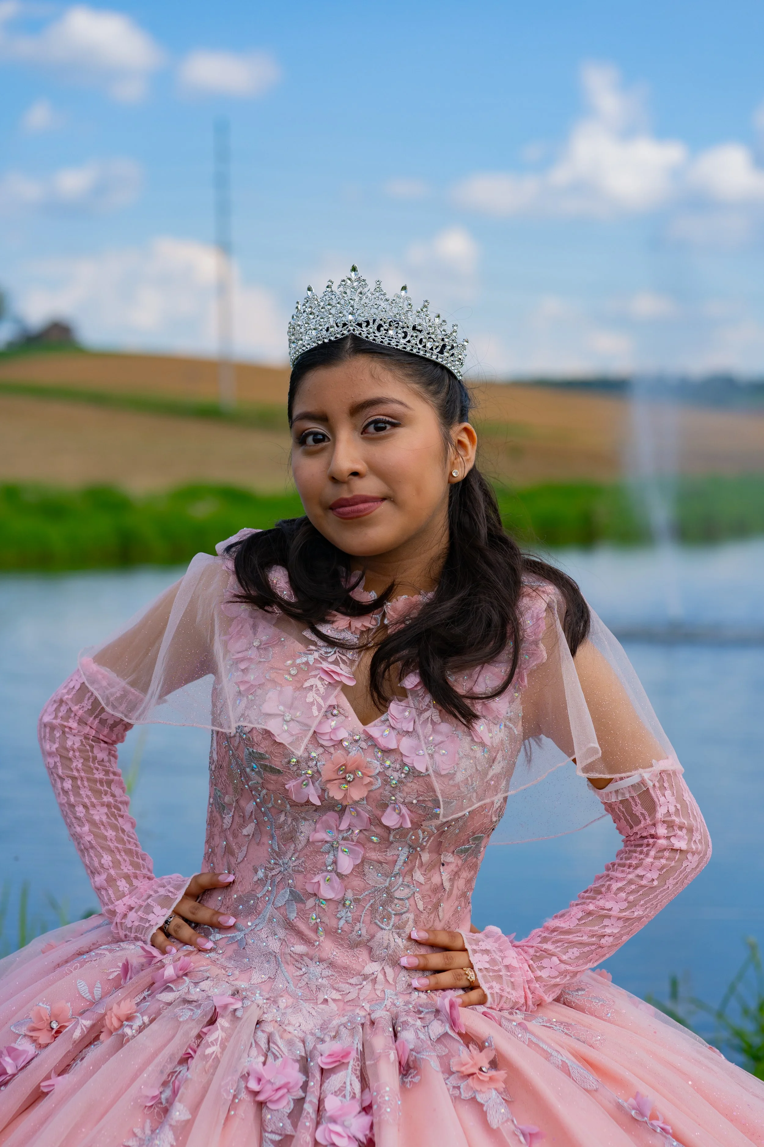 A young woman wearing a pink princess gown with lace and floral embellishments, and a silver tiara, posing outdoors near a body of water with green grass and rolling hills in the background.
