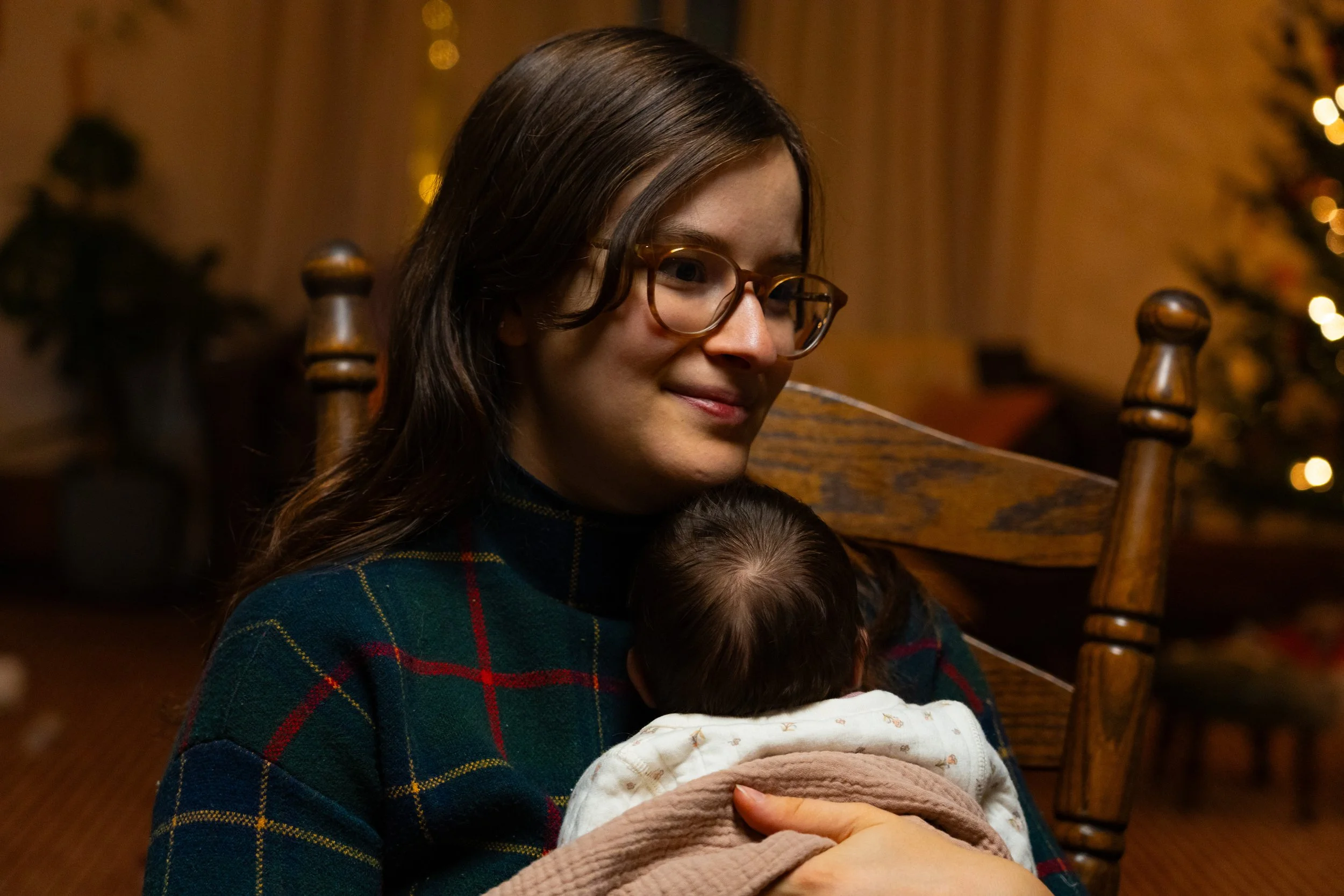 A woman with long brown hair, glasses, and a tartan shirt, smiling gently while holding a sleeping baby wrapped in a blanket. The background features warm lighting and festive decorations.