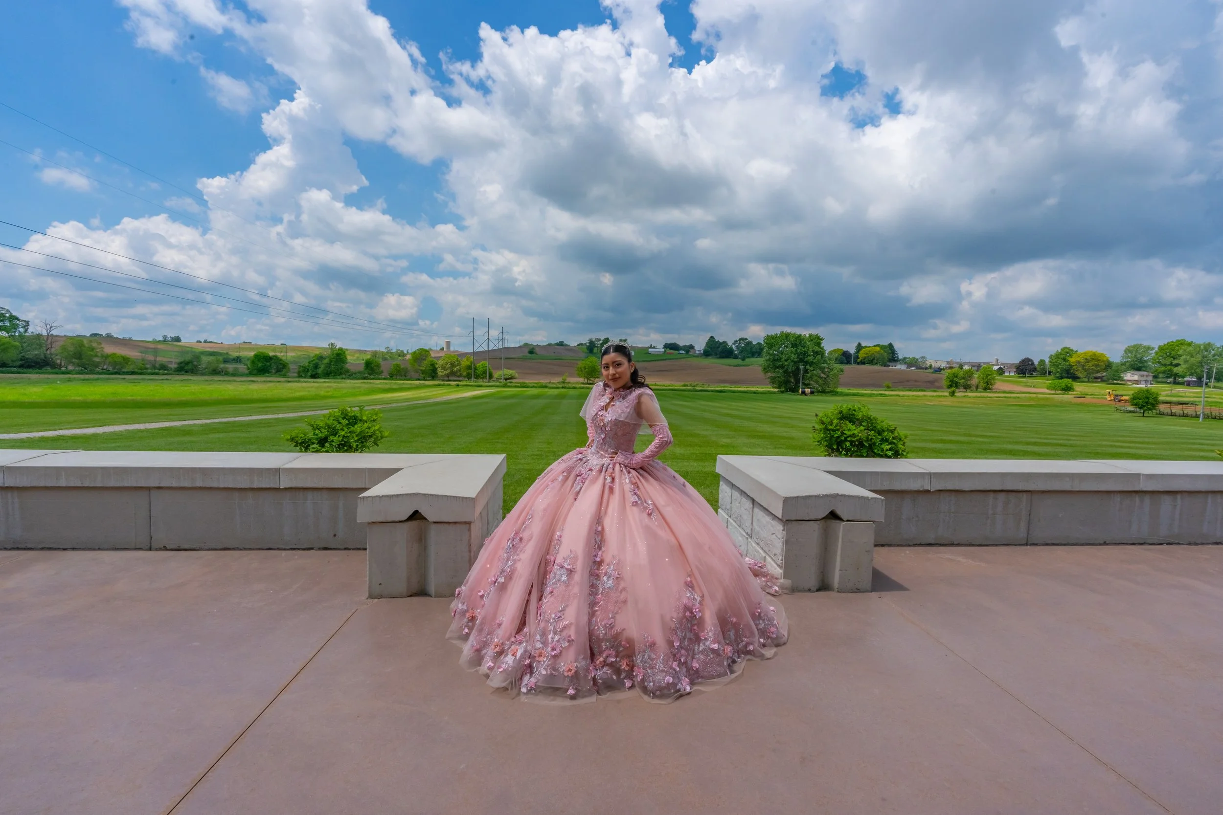 A woman in an elegant pink gown with floral embellishments standing outdoors on a stone terrace with green landscape and blue sky with clouds in the background.
