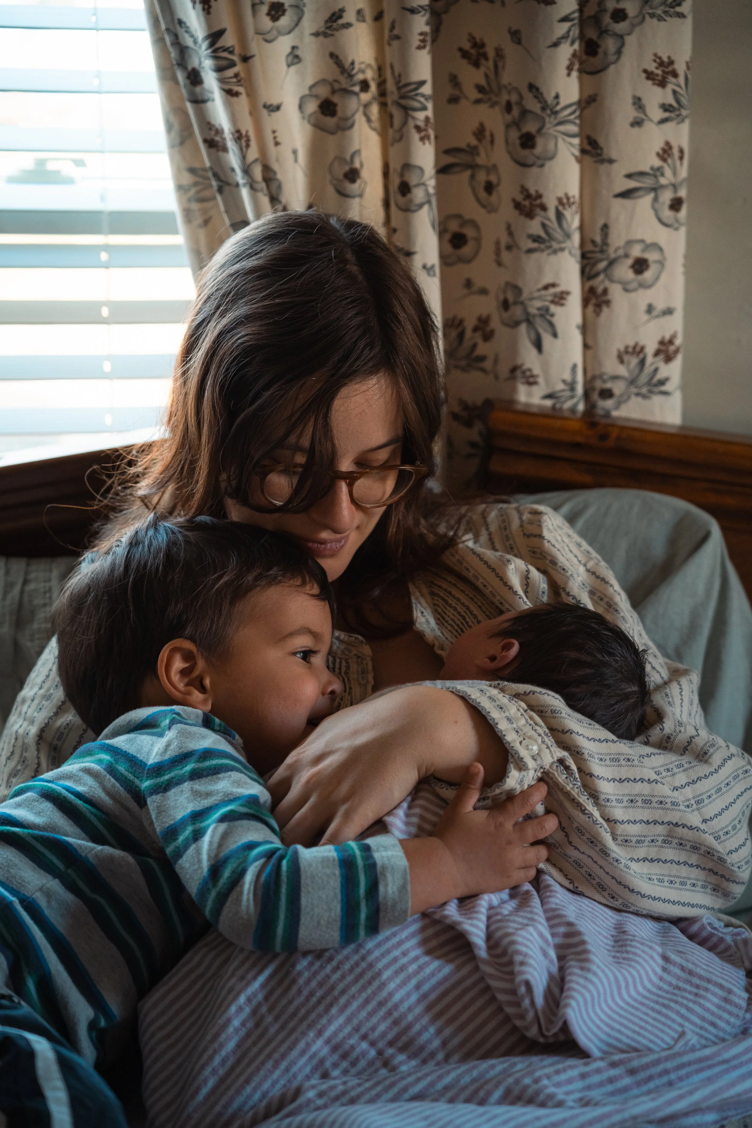 A woman with brown hair and glasses cuddling with three young children in a cozy room with floral curtains, sunlight coming through blinds, and a wooden headboard.
