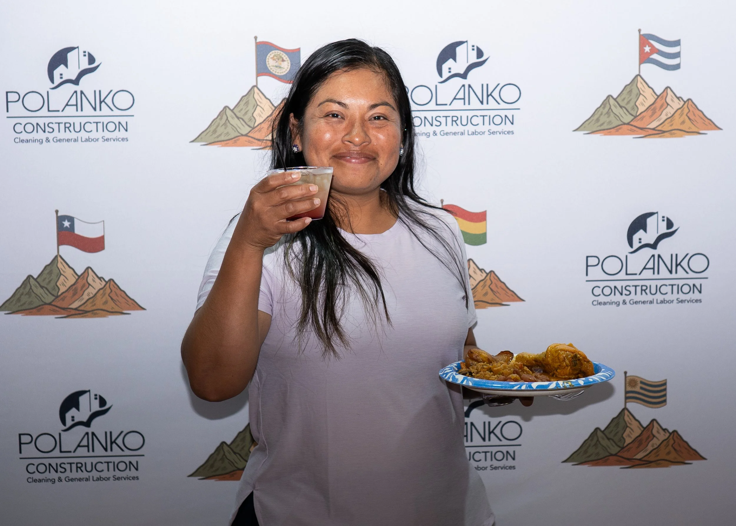 A woman with dark hair and earrings smiling while holding a plate of fried chicken and a clear cup of drink in front of a backdrop with the logo and mountains of Polanko Construction.