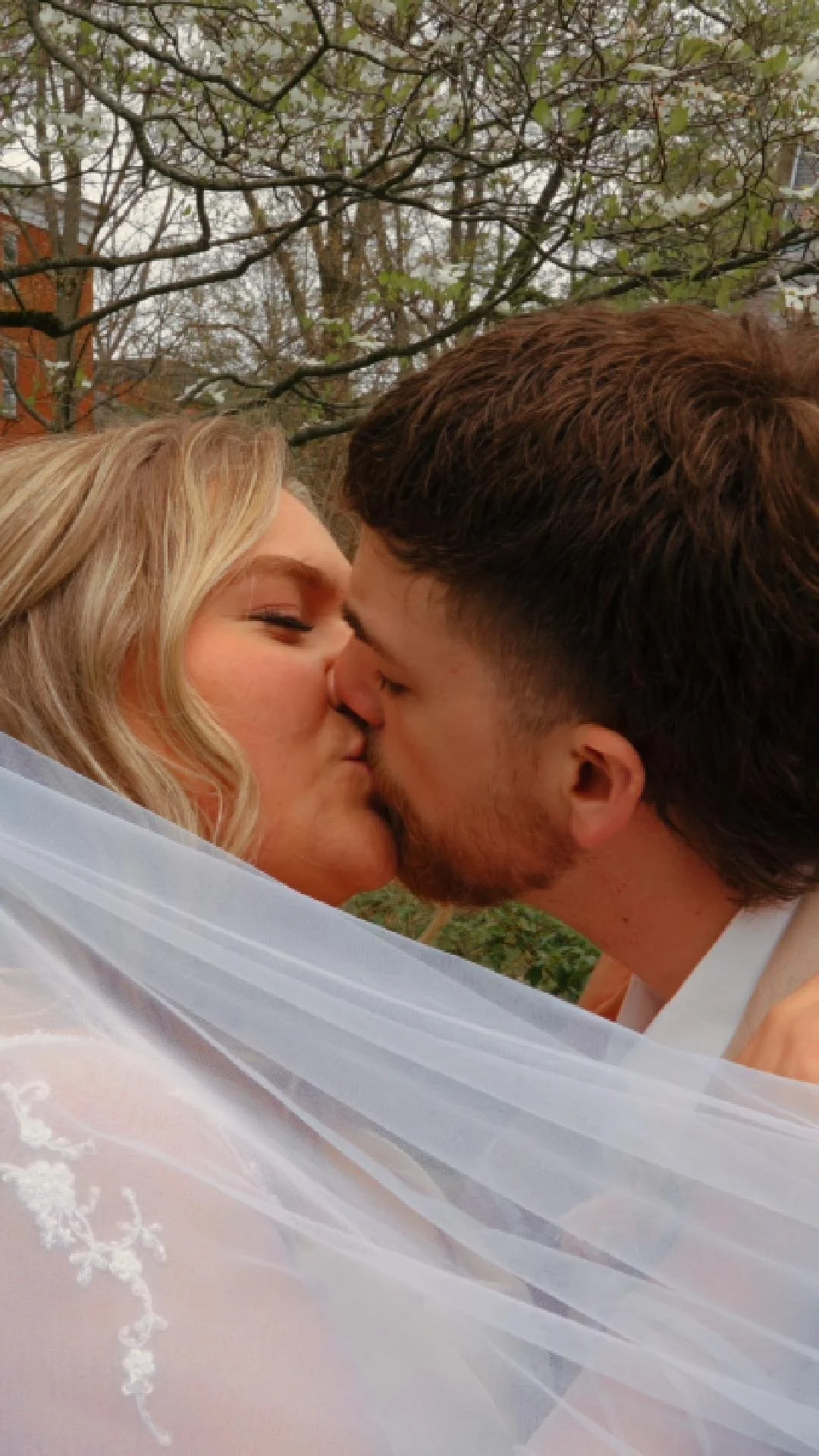 Couple kissing outdoors, with trees in the background, a woman with blonde hair and a man with brown hair.