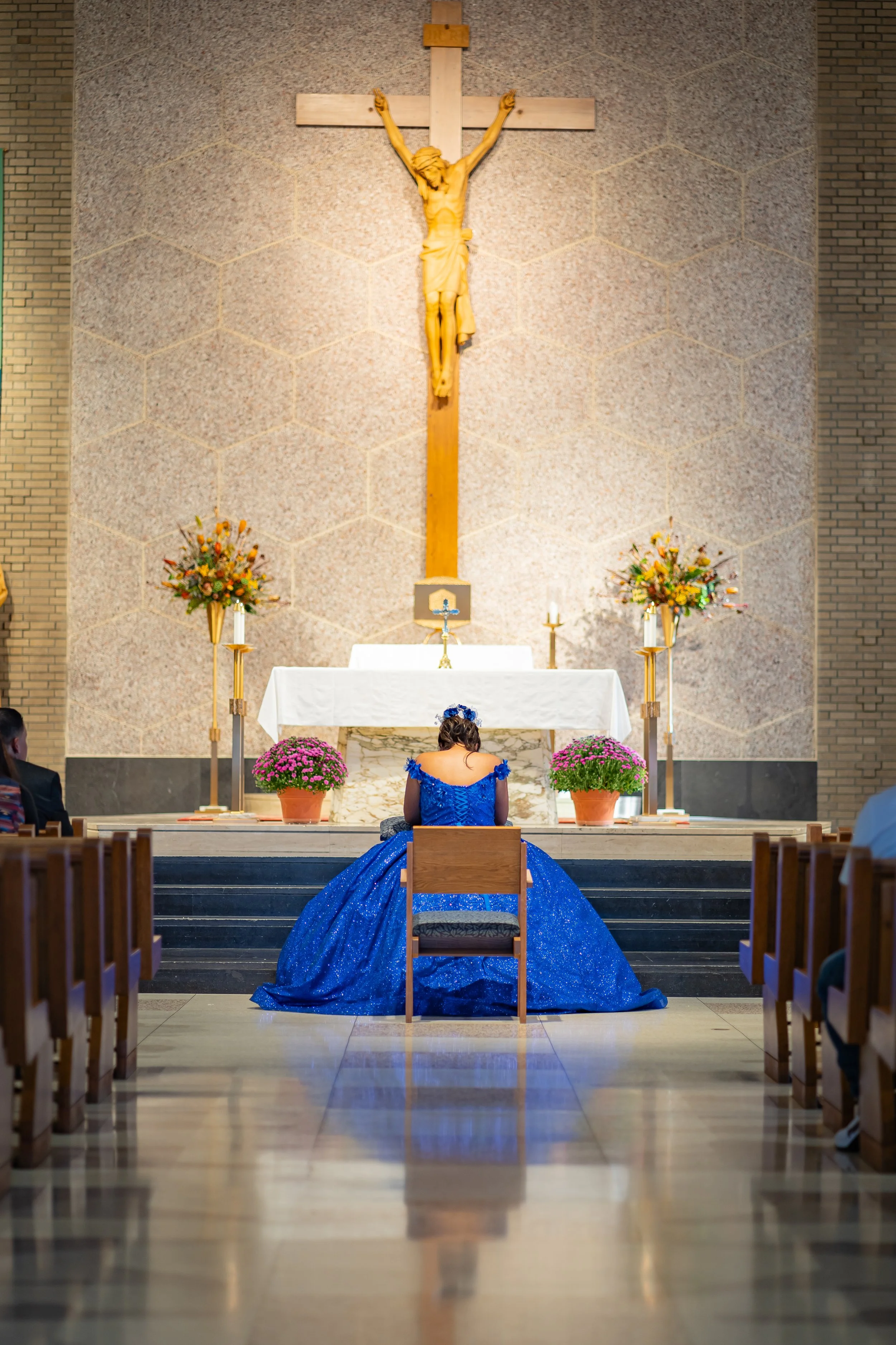A woman in a blue dress kneels in prayer at the altar of a church, with a crucifix on the wall and flowers surrounding her.