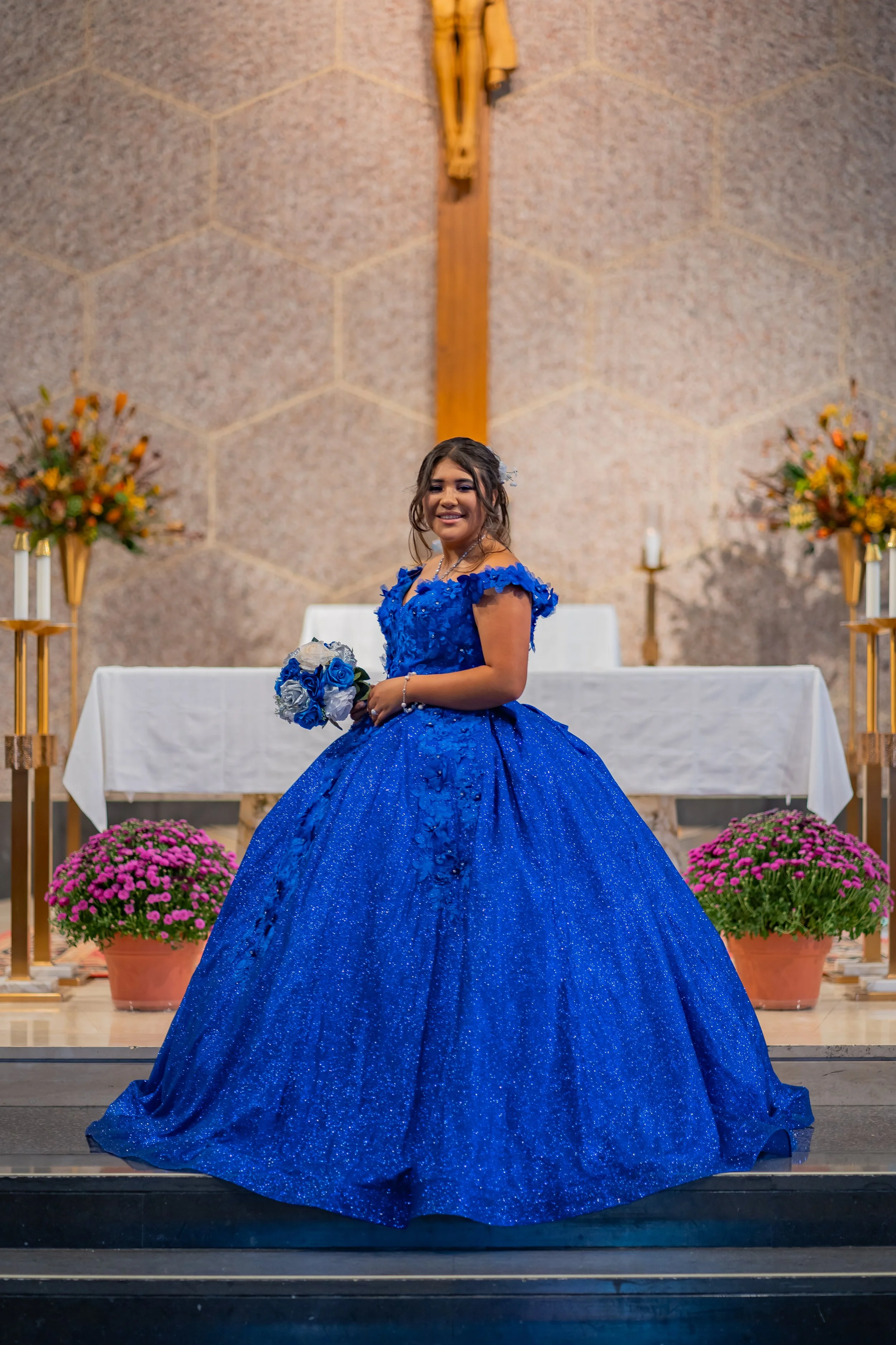 A young woman in a vibrant blue, ball gown-style dress standing in front of an altar in a church. She is holding a bouquet of blue and white flowers and smiling at the camera. The background features a cross on the wall, floral arrangements, and cand