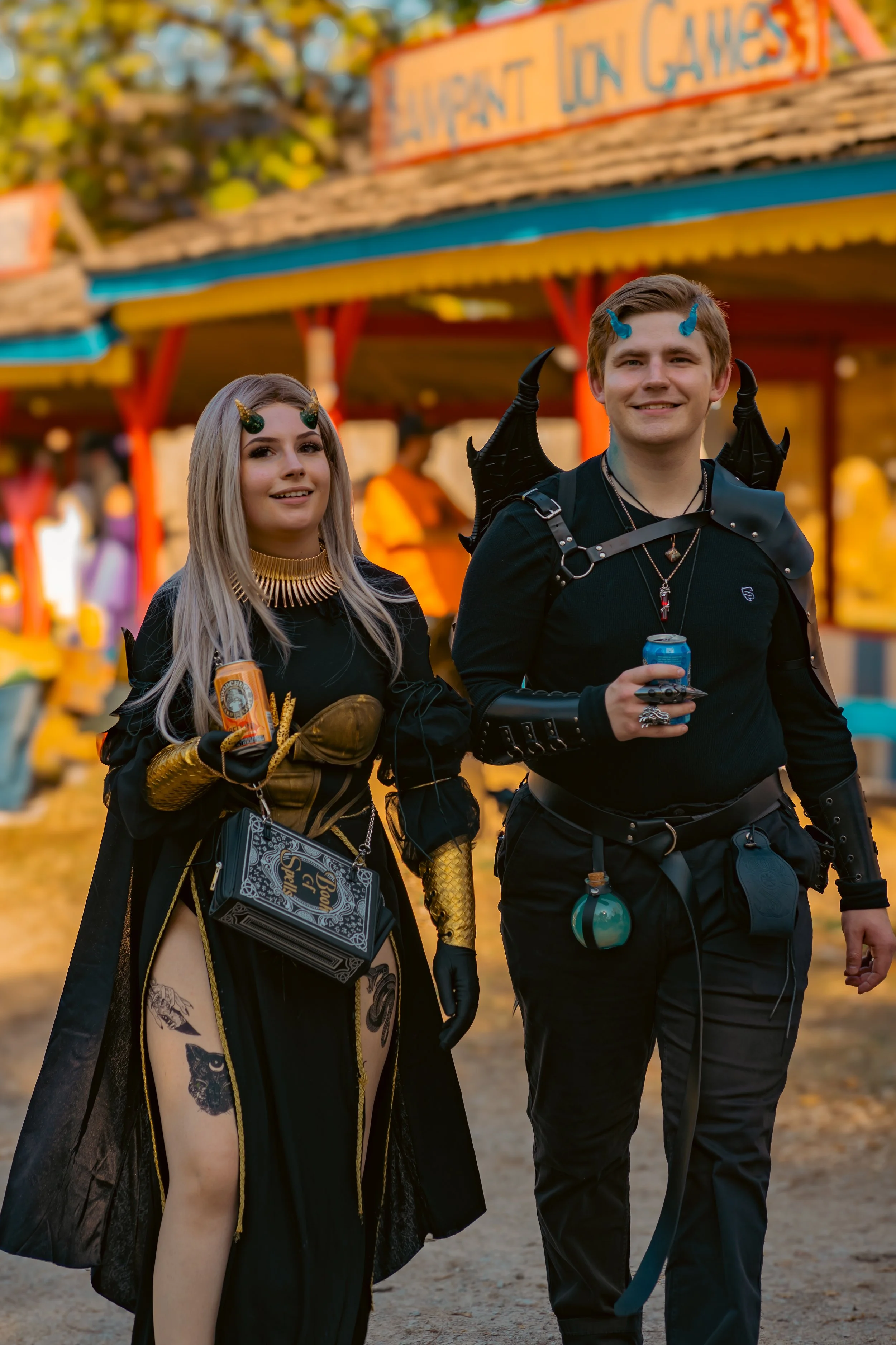 Two individuals dressed in fantasy or cosplay costumes walking outdoors at what appears to be a fair or carnival. The woman has long, blonde hair, small black horns, and tattoos, and is holding a can. The man has short brown hair, small blue horns, a