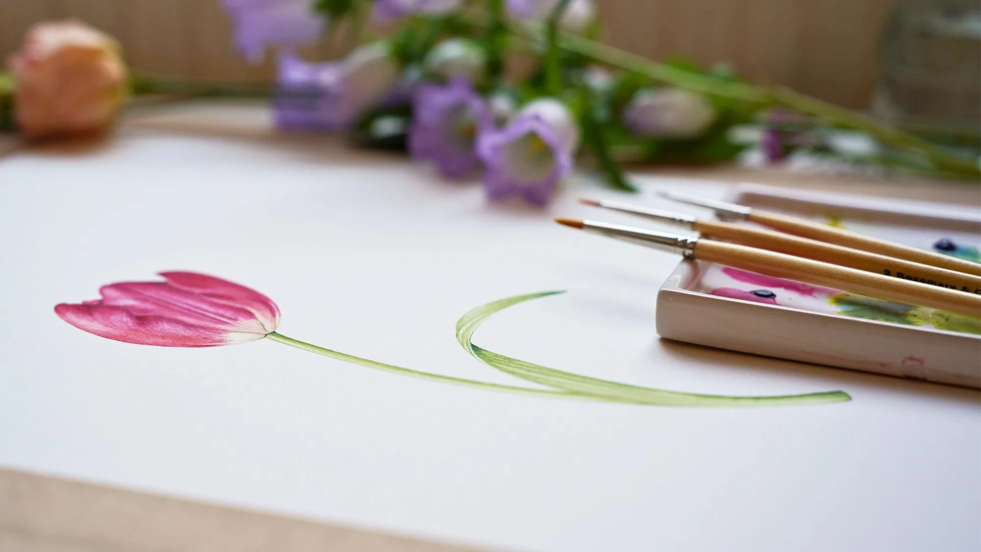 Watercolour painting of a pink tulip with brushes and palette on the table, demonstrating botanical illustration for beginners.