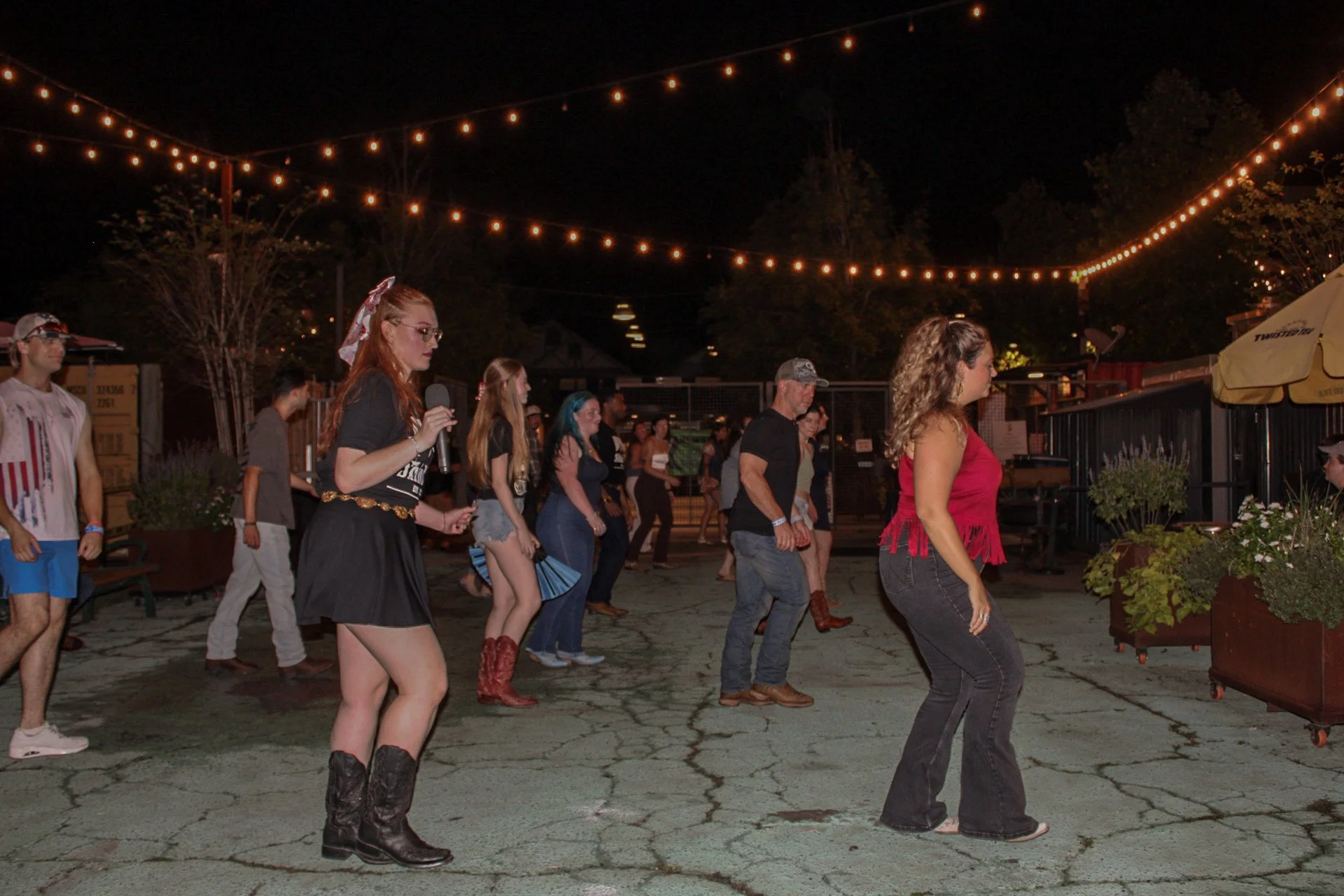 People line dance outdoors at night under string lights with trees and a patio in the background.