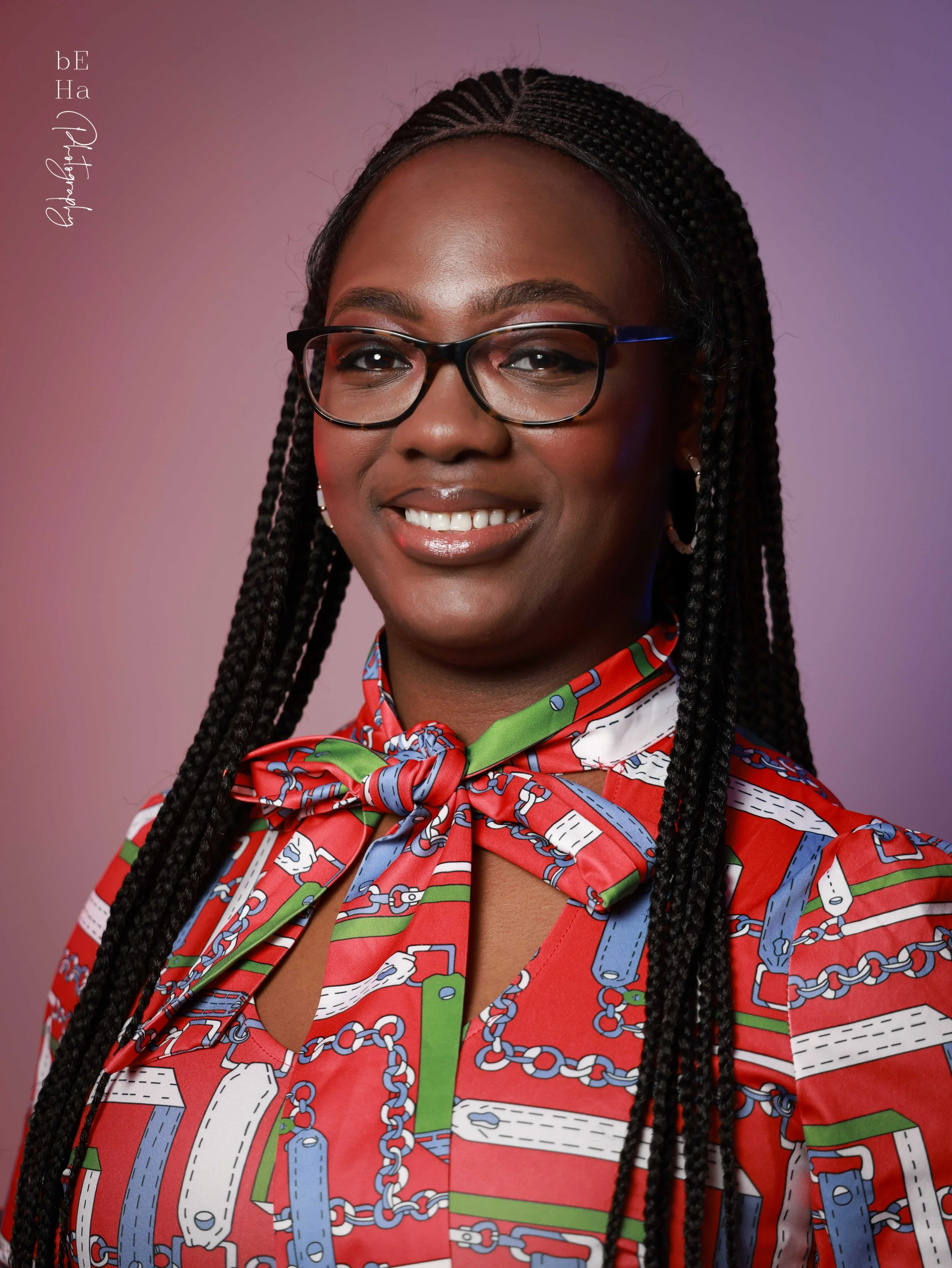 Close-up of a woman with dark hair in braids, wearing glasses, smiling, and dressed in a colorful patterned shirt with a scarf tied at the neck. The background is gradient purple.