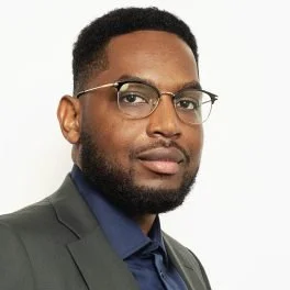 Portrait of a man wearing glasses, a dark blazer, and a blue shirt against a plain white background.