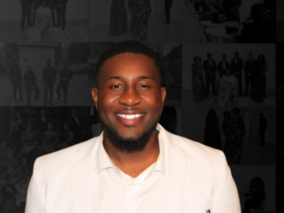A smiling man with a beard and short hair, wearing a white blazer, against a dark collage background.