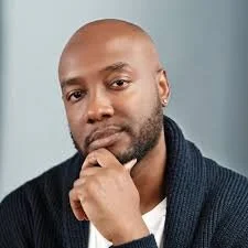A man with a beard and bald head, wearing a dark sweater, posing with his hand on his chin against a neutral background.