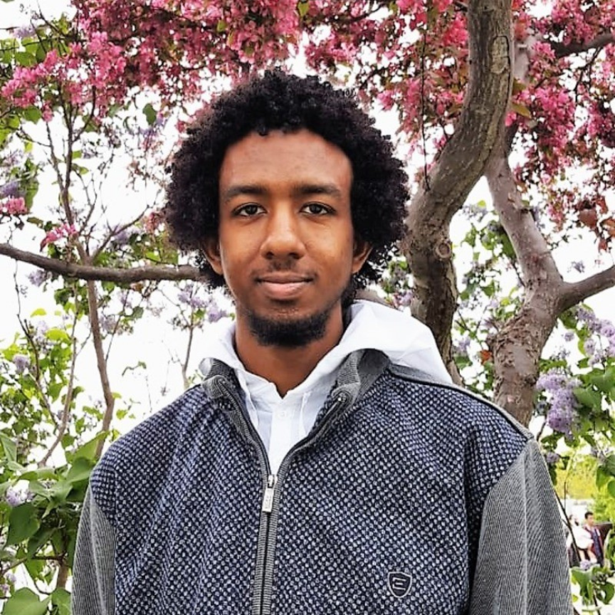 A young man with curly black hair and a slight beard is standing outdoors in front of a flowering tree with pink blossoms. He is wearing a gray jacket over a white hoodie, and the background shows more trees and green foliage.