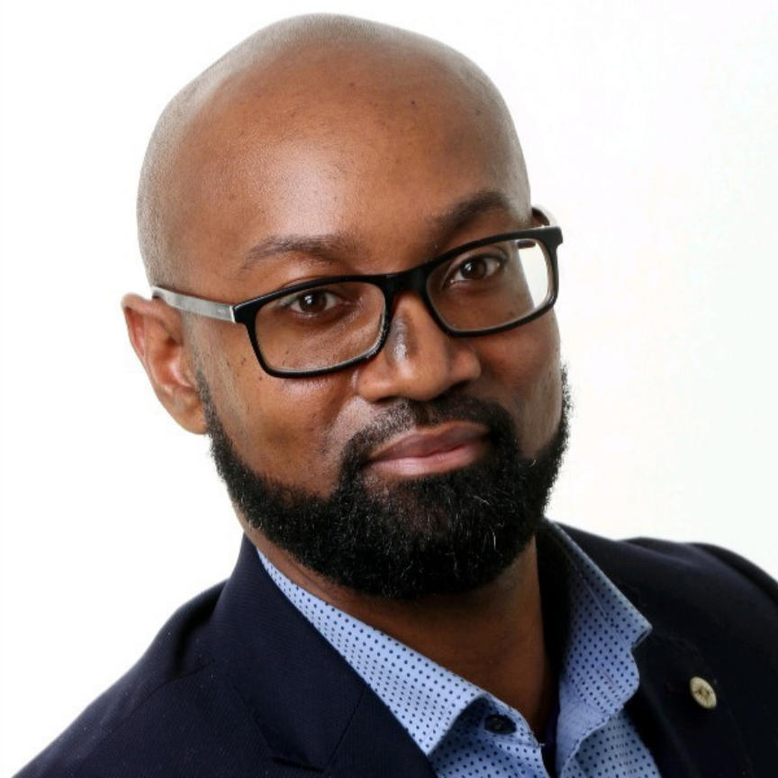 Portrait of a bald man with glasses, a beard, dressed in a dark blazer and a light blue collared shirt, against a white background.