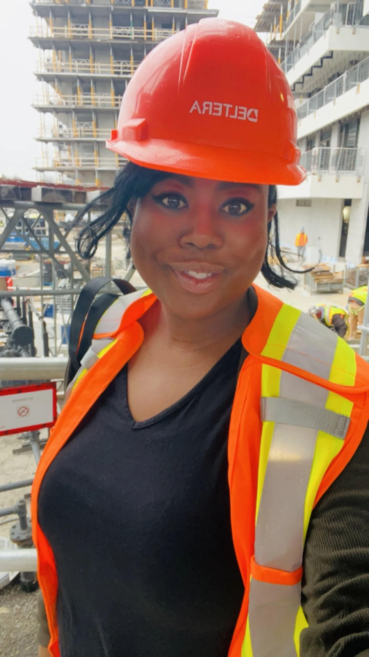 A woman wearing a red construction helmet with the word 'DELETERRA' on it, a high-visibility safety vest, and a black shirt, standing at a construction site with a partially built high-rise building in the background.