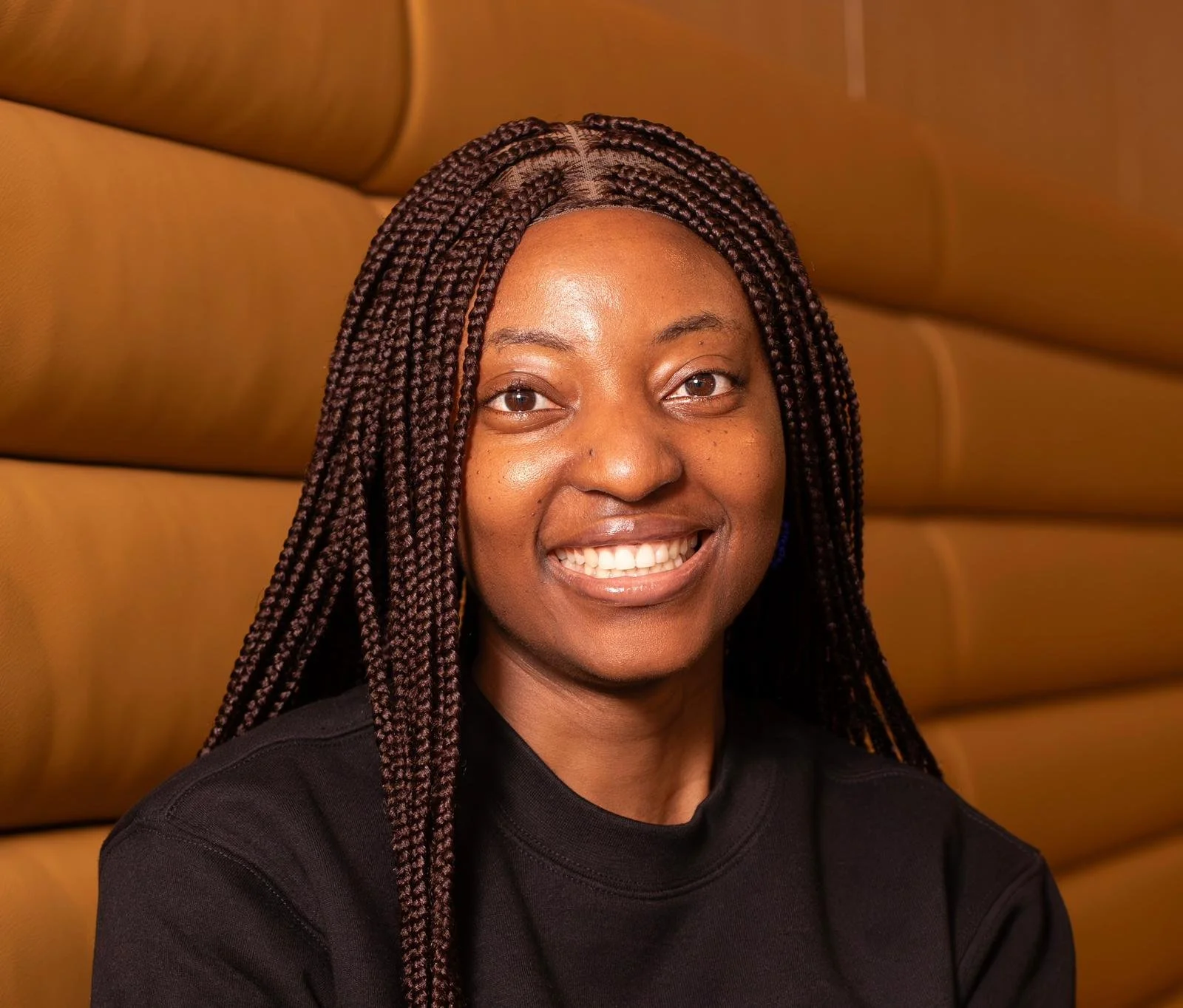 A woman with braided hair and a bright smile sitting against a brown cushioned wall.