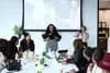 A diverse group of women in a professional setting attending a meeting or presentation, with a woman standing and speaking at a conference table, some taking photos, in a bright modern office.