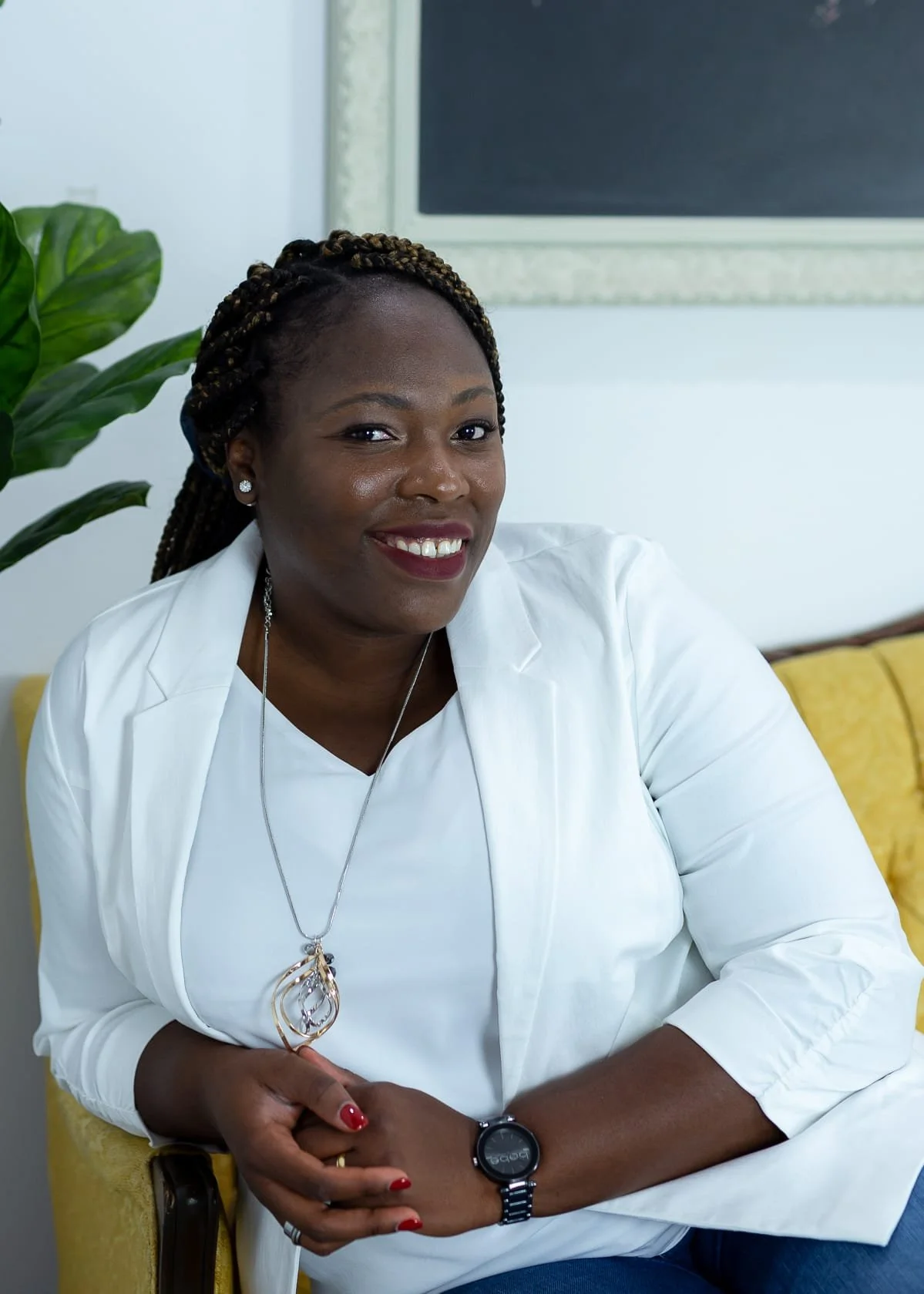 A smiling African American woman with braided hair, wearing a white blazer and jewelry, sitting on a yellow chair indoors, with a large green plant to her left and a blackboard behind her.