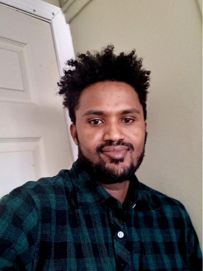 A man with curly hair, facial hair, and dark skin taking a selfie indoors, wearing a green and black plaid shirt.